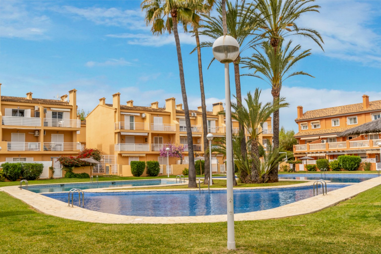 Vista de la piscina comunitaria con acceso escalonado, rodeada de césped y edificios residenciales color melocotón.