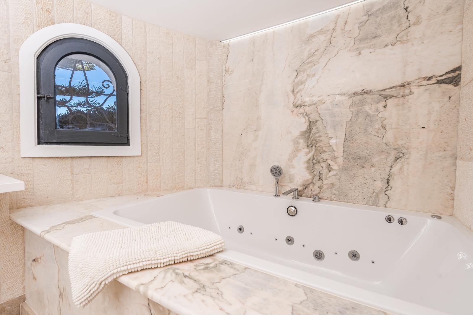 Bañera de hidromasaje integrada con revestimiento de mármol veteado y paredes de piedra natural. Incluye una ventana de arco mediterránea y retroiluminación LED superior.
