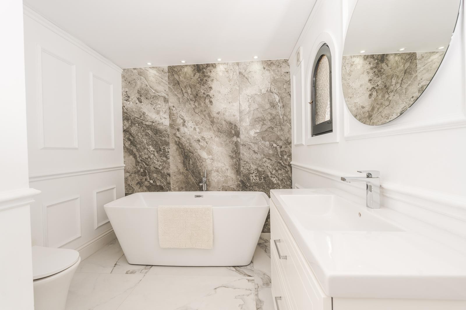 Baño de la suite principal con bañera exenta, pared de mármol veteado y molduras decorativas. Incluye lavabo moderno, espejo circular y una ventana de arco de estilo mediterráneo.