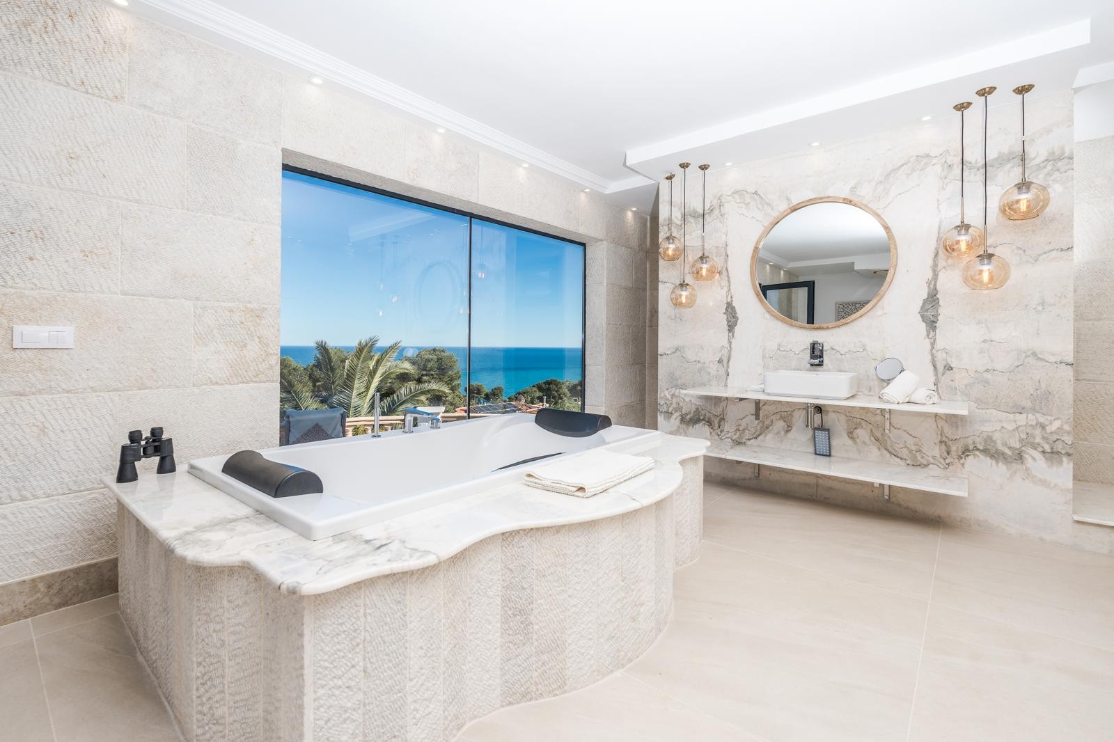 Baño de la suite principal con bañera frente a un gran ventanal con vistas al mar, paredes de mármol veteado, estantes flotantes y elegante iluminación colgante.