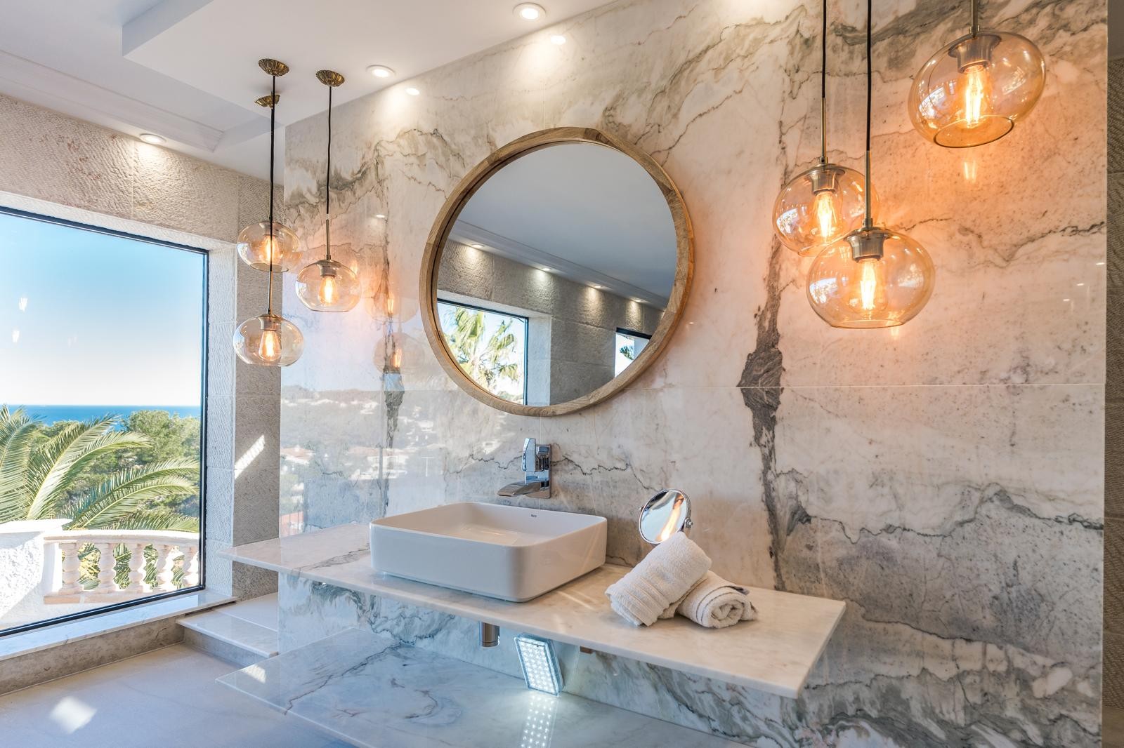 Baño de la suite principal con paredes de mármol veteado, lavabo sobre encimera, espejo circular de madera y vistas panorámicas al mar a través del gran ventanal.