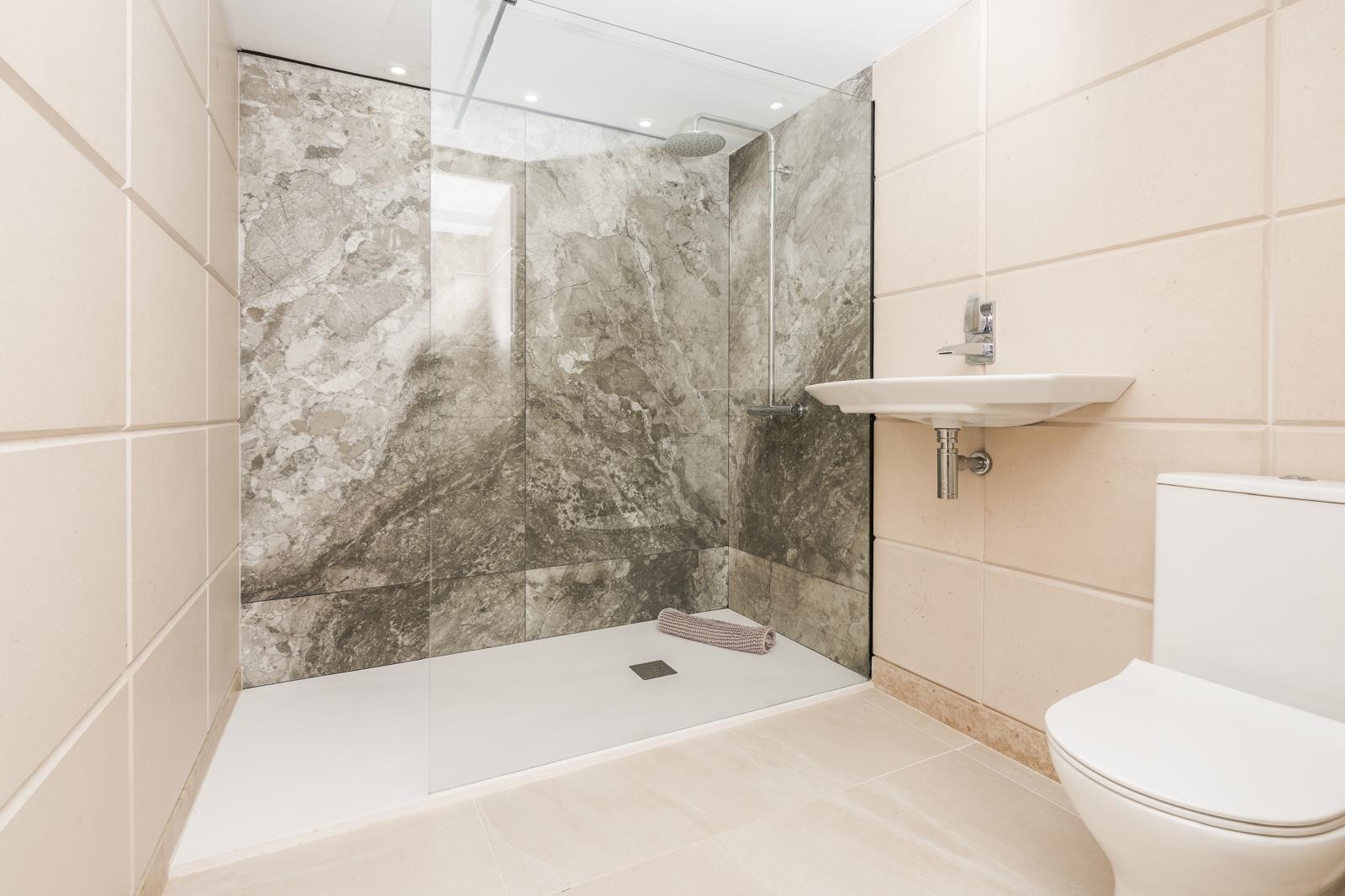 Baño moderno con ducha walk-in revestida en mármol gris, mampara de cristal fija y sanitarios blancos integrados en paredes de tonos neutros.