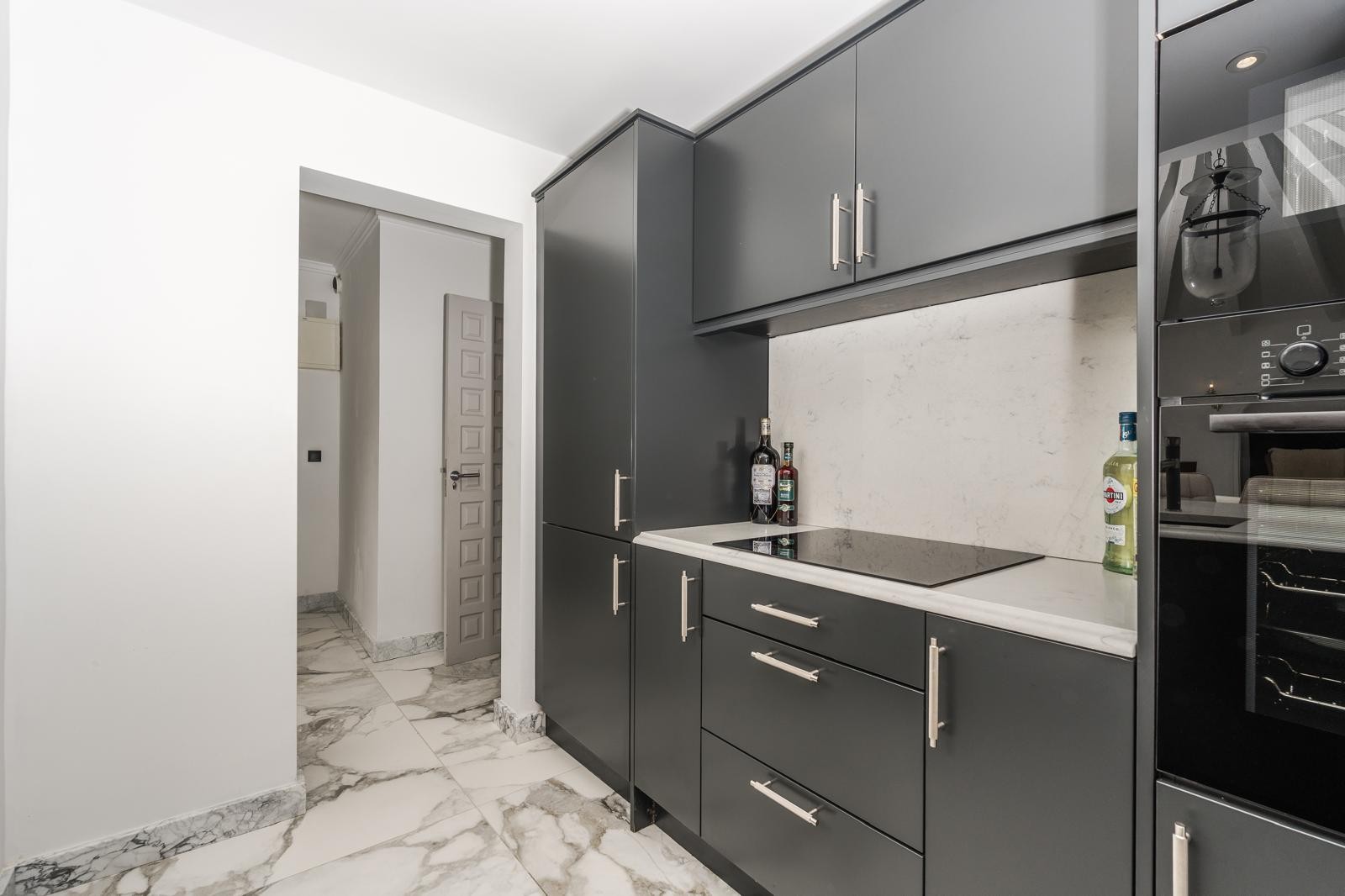 Cocina contemporánea con armarios gris oscuro, encimera de mármol blanco y placa de inducción integrada sobre un suelo de mármol veteado.