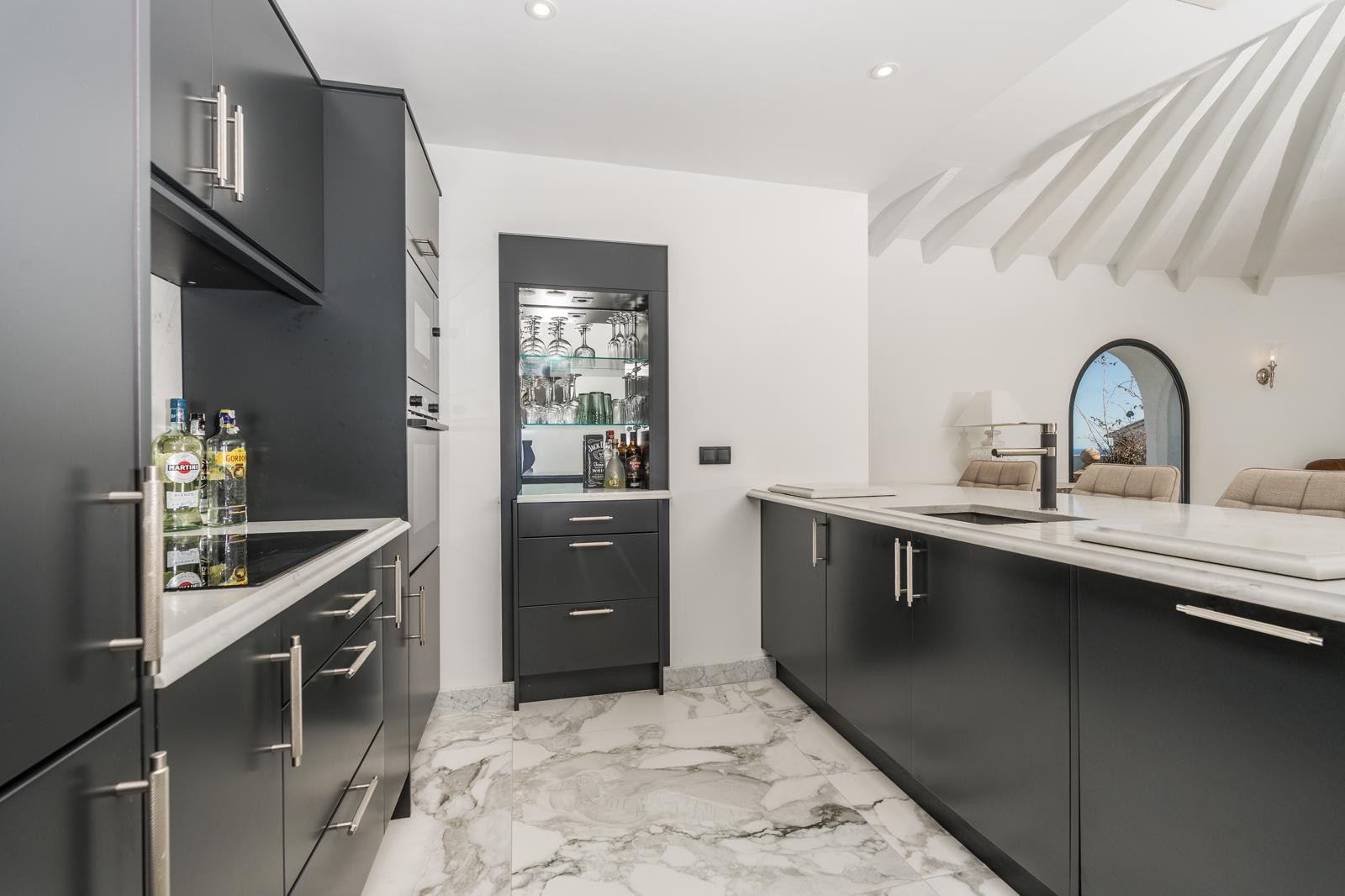 Cocina moderna con armarios negro mate, encimeras de mármol blanco y suelo coordinado. Incluye una vitrina integrada y vistas al techo de vigas blancas del salón circular adyacente.