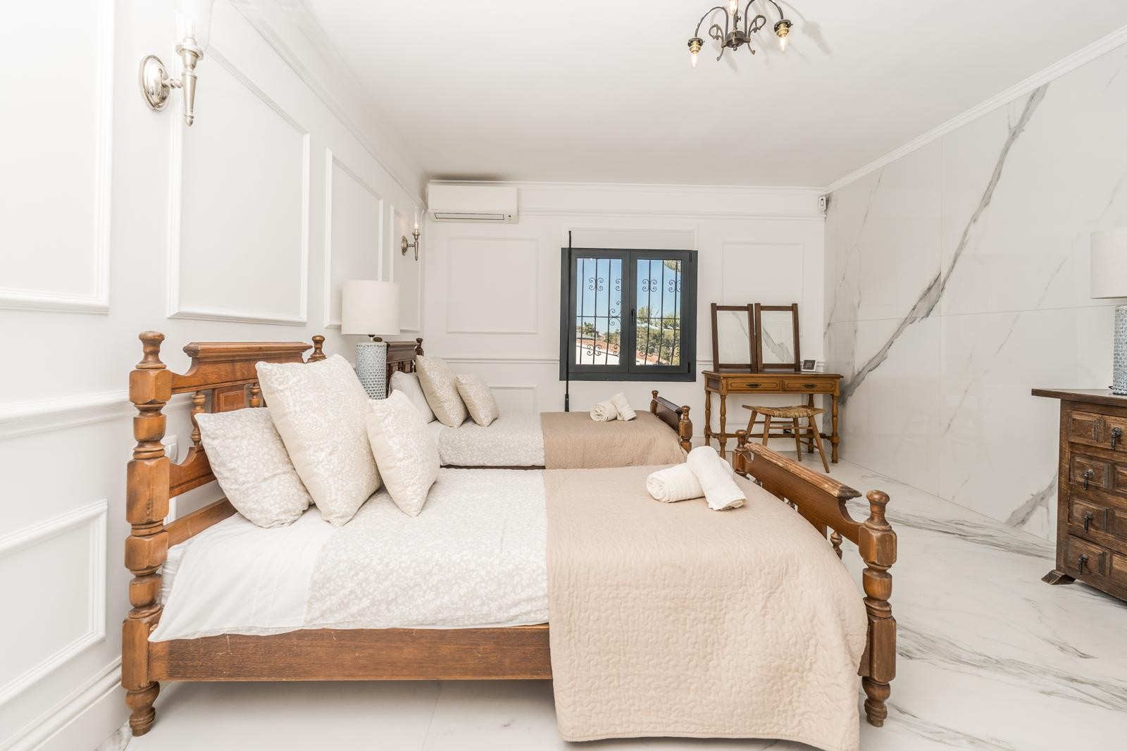 Dormitorio con dos camas de madera, paredes con molduras clásicas y suelos de mármol. Incluye un tocador rústico, aire acondicionado y una pared de acento en piedra natural.
