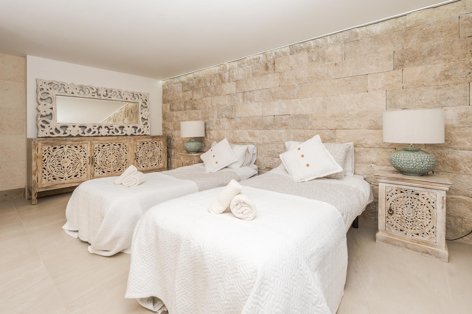 Dormitorio con dos camas individuales destacando una pared de piedra natural, mobiliario de madera tallada artesanalmente y suelos de gres en tonos neutros.