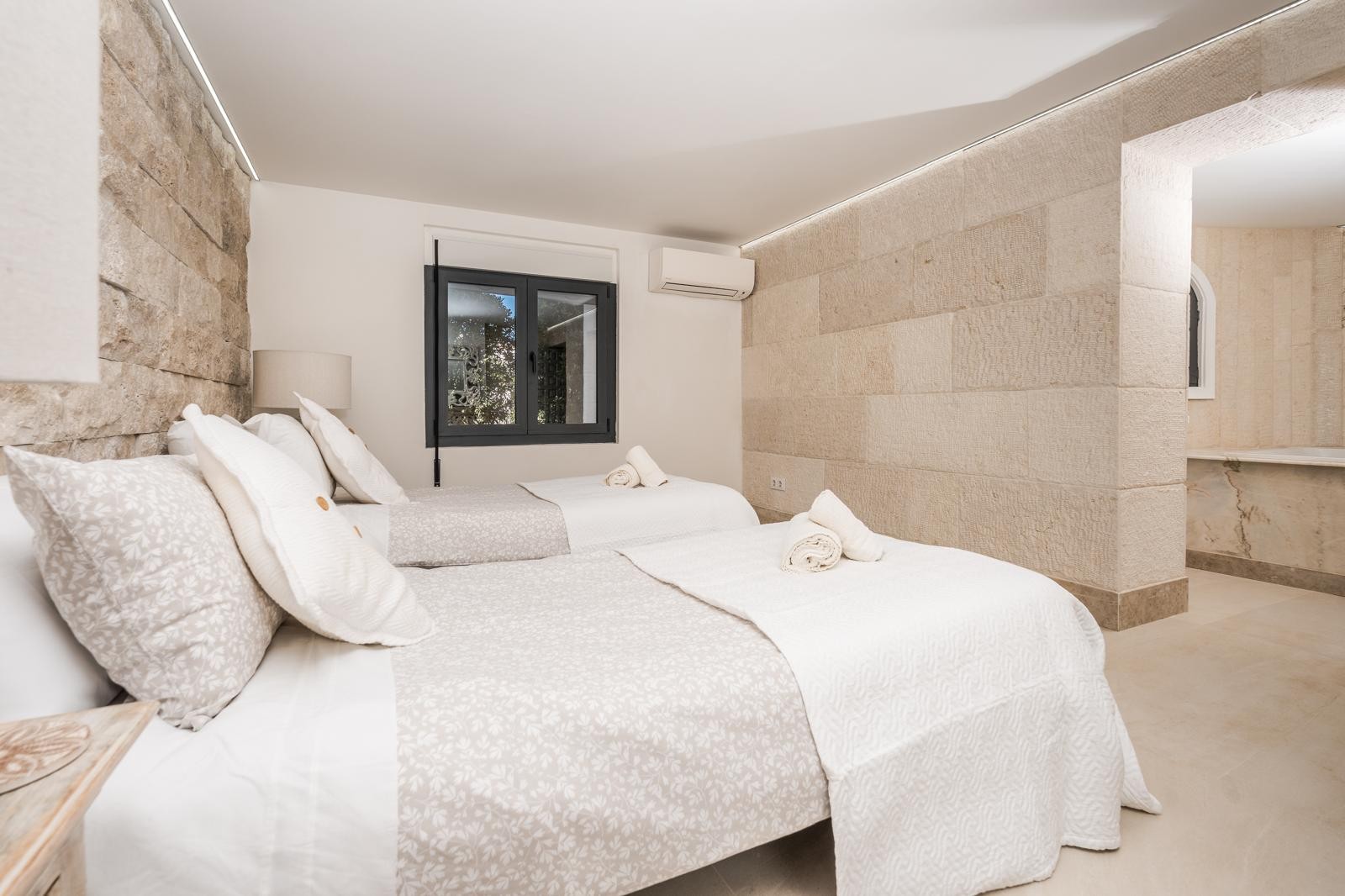 Dormitorio con paredes de piedra natural, dos camas individuales, aire acondicionado y acceso a un baño integrado con acabados en mármol.