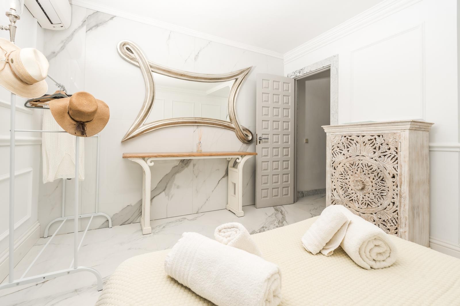 Dormitorio con revestimiento de mármol veteado, un gran espejo ornamental de marco plateado y una cómoda de madera tallada de estilo artesanal.