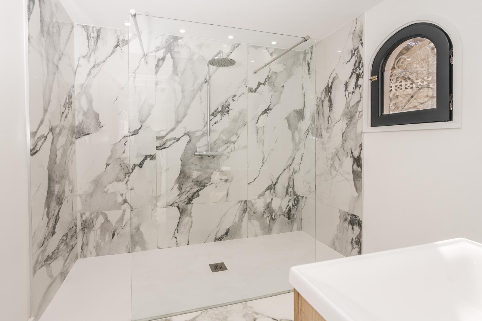 Ducha italiana con revestimiento de mármol de gran formato, mampara de cristal fija, grifería cromada y ventana de arco con marco negro.