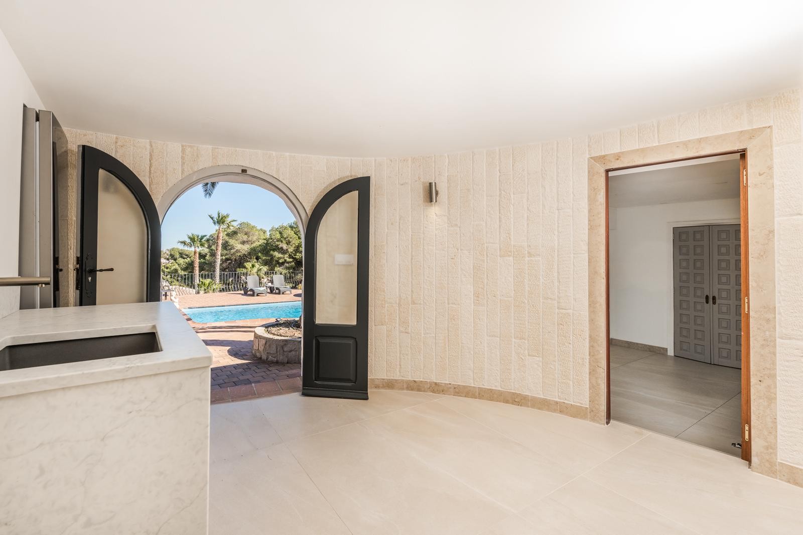 Espacio interior con paredes revestidas de piedra natural y encimera de mármol, con acceso directo a la zona de piscina a través de un gran ventanal de arco.