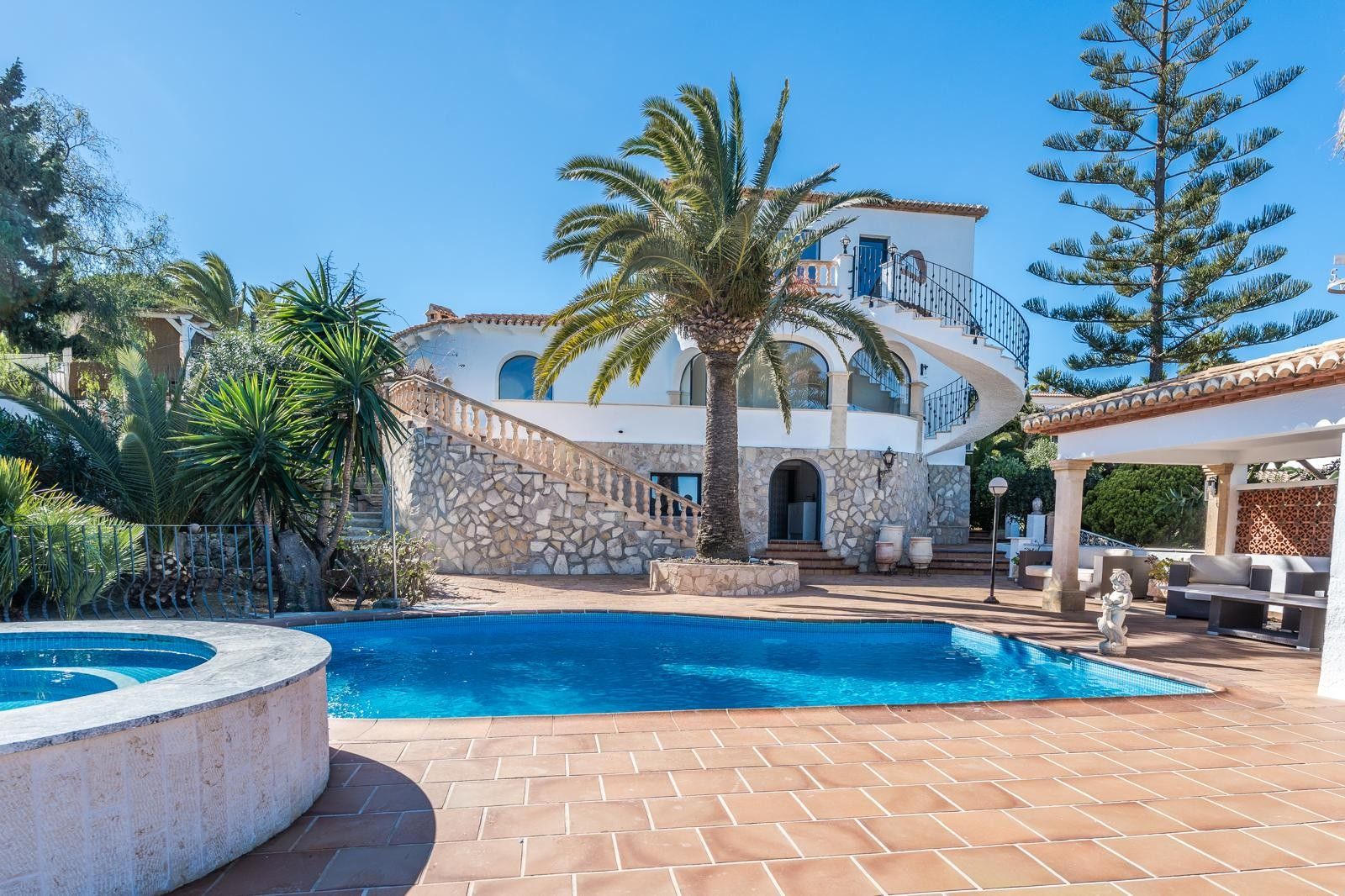 Exterior de la villa con piscina, jacuzzi circular y fachada de piedra natural. Destaca la escalera de caracol exterior y el jardín mediterráneo con palmeras.