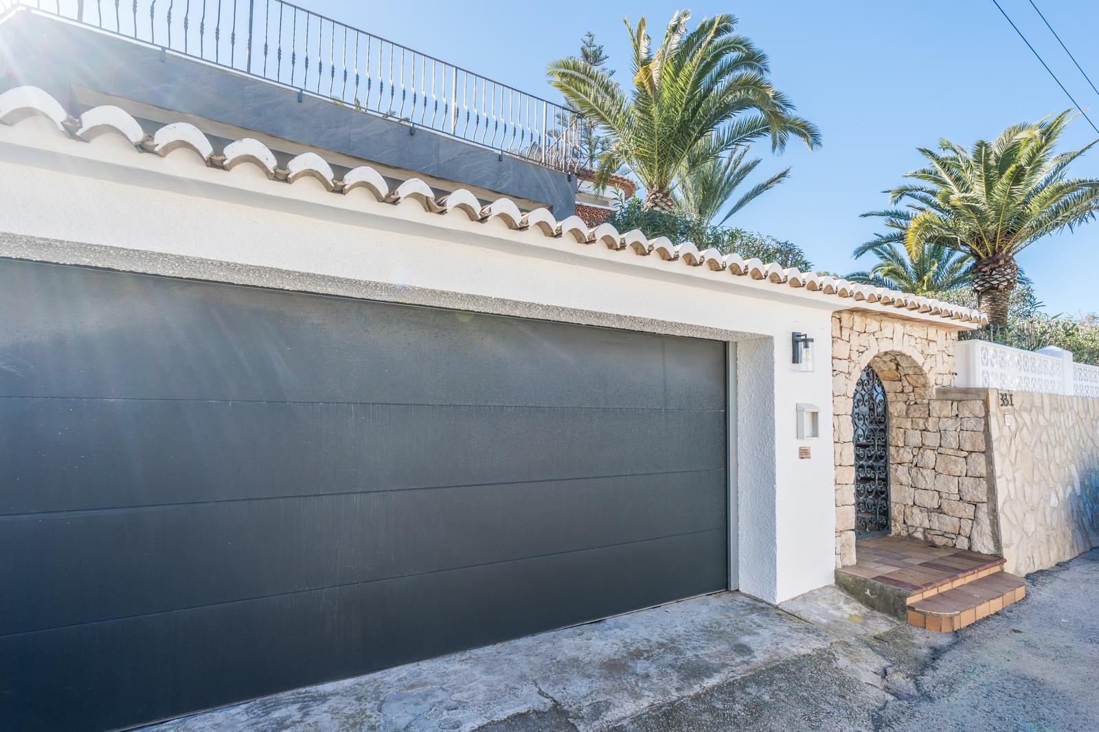 Fachada exterior con garaje doble de puerta oscura, arco de piedra natural con puerta de forja y muro perimetral de piedra seca bajo palmeras mediterráneas.