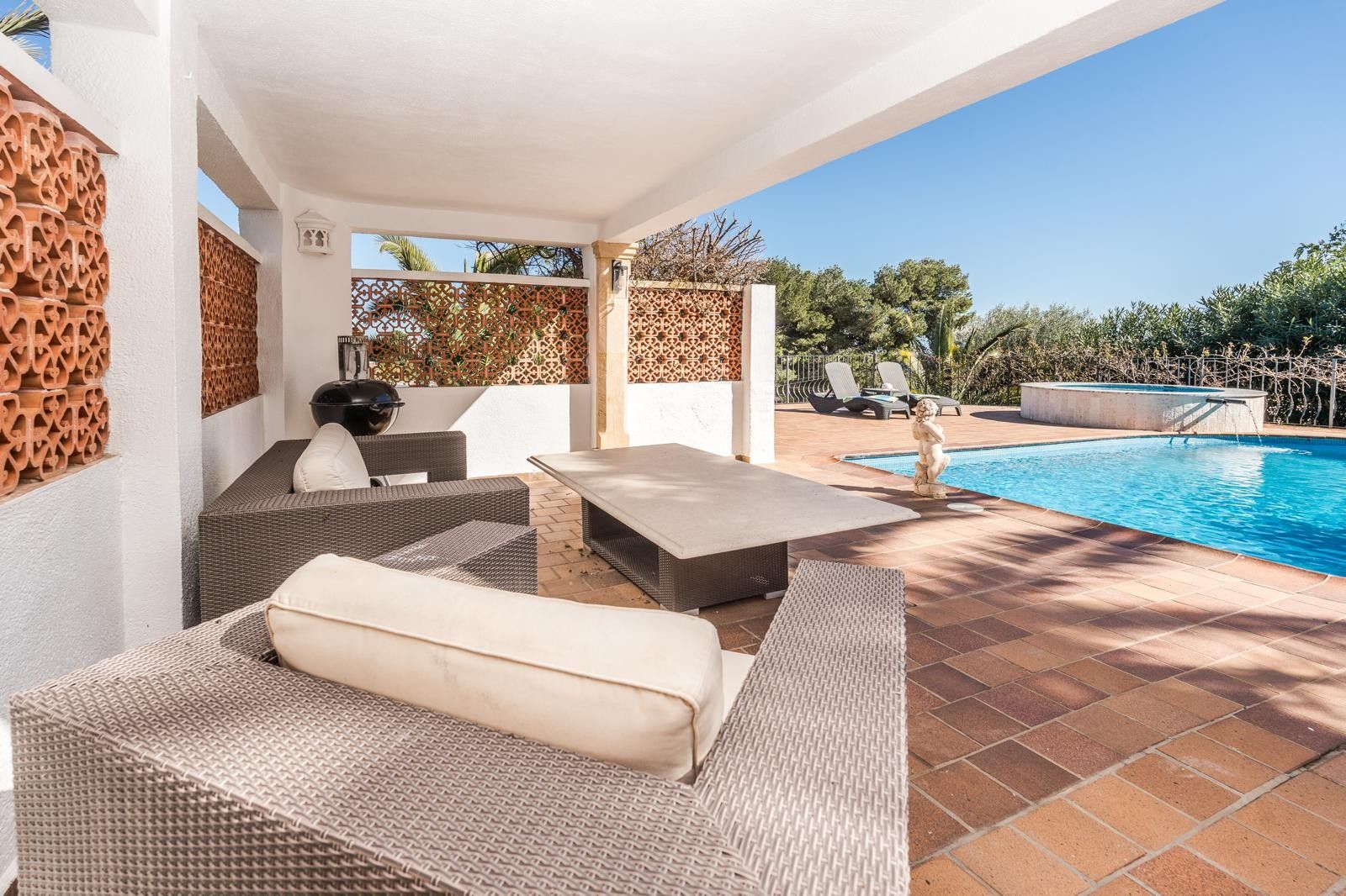 Porche cubierto con mobiliario de ratán y celosías de terracota, situado frente a la piscina renovada y el jacuzzi circular rodeado de vegetación.