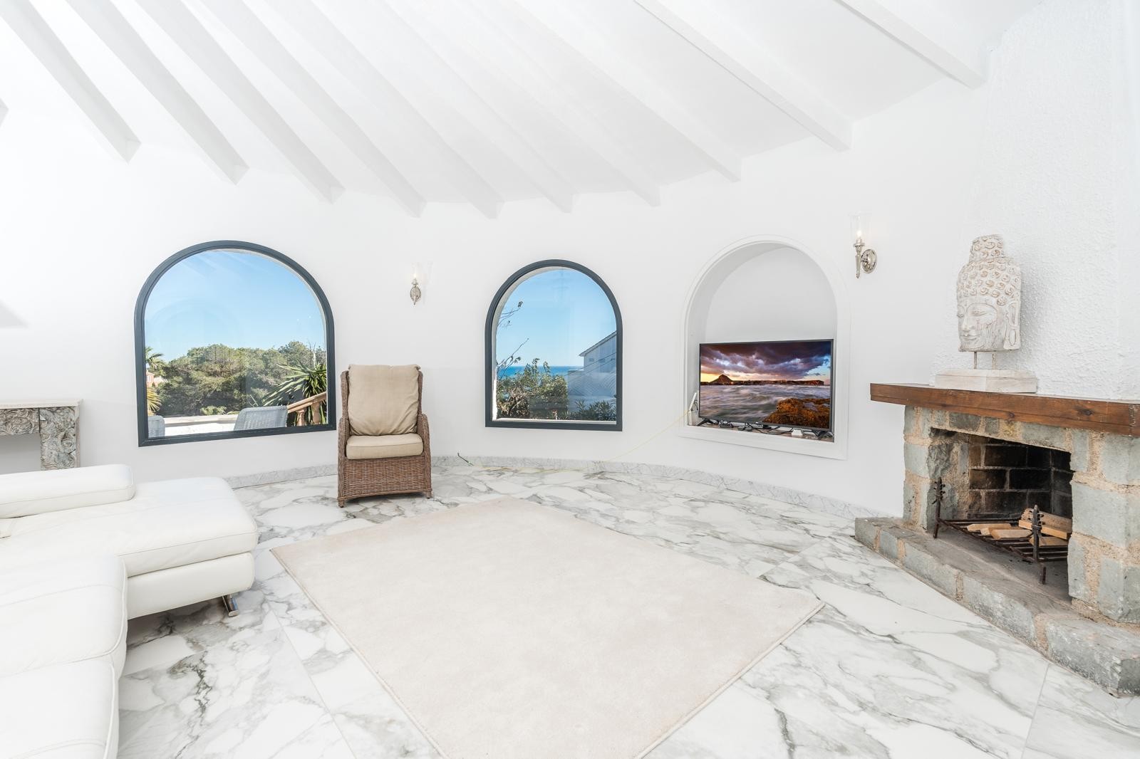 Salón circular con techos de vigas blancas, suelo de mármol y chimenea de piedra natural. Ventanales de medio punto ofrecen vistas al entorno mediterráneo y al mar.