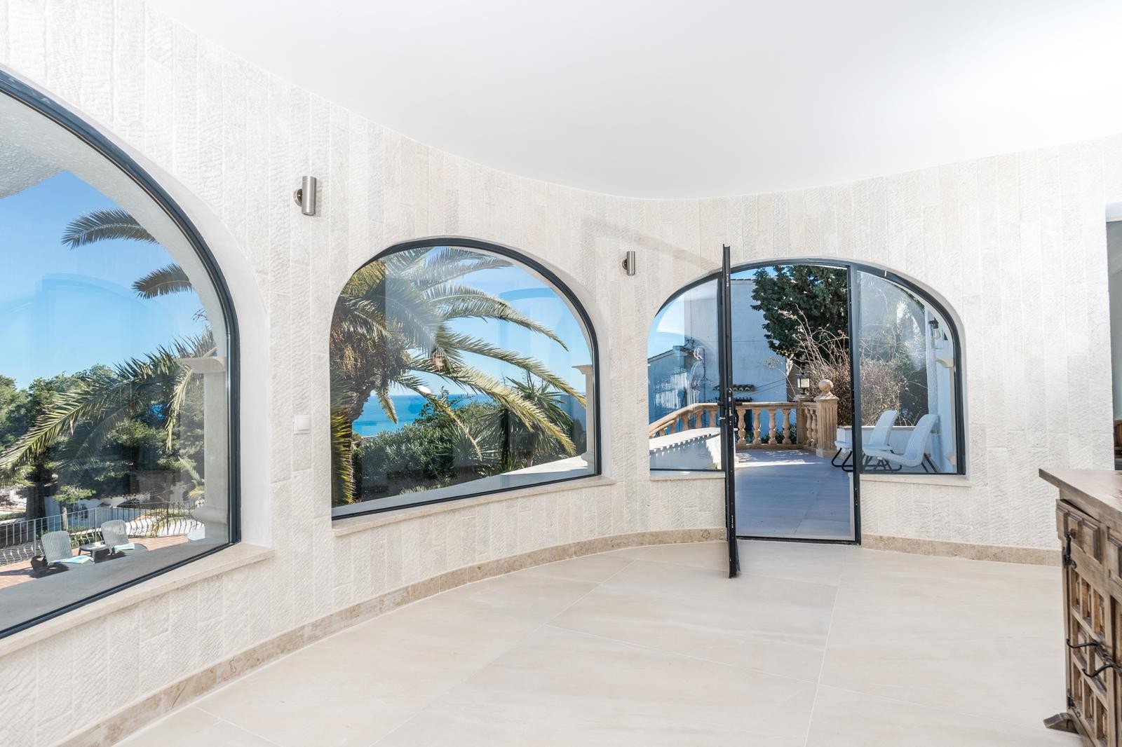 Salón de planta circular con muros revestidos en piedra natural y amplios ventanales en arco que ofrecen vistas al mar y acceso directo a la terraza.
