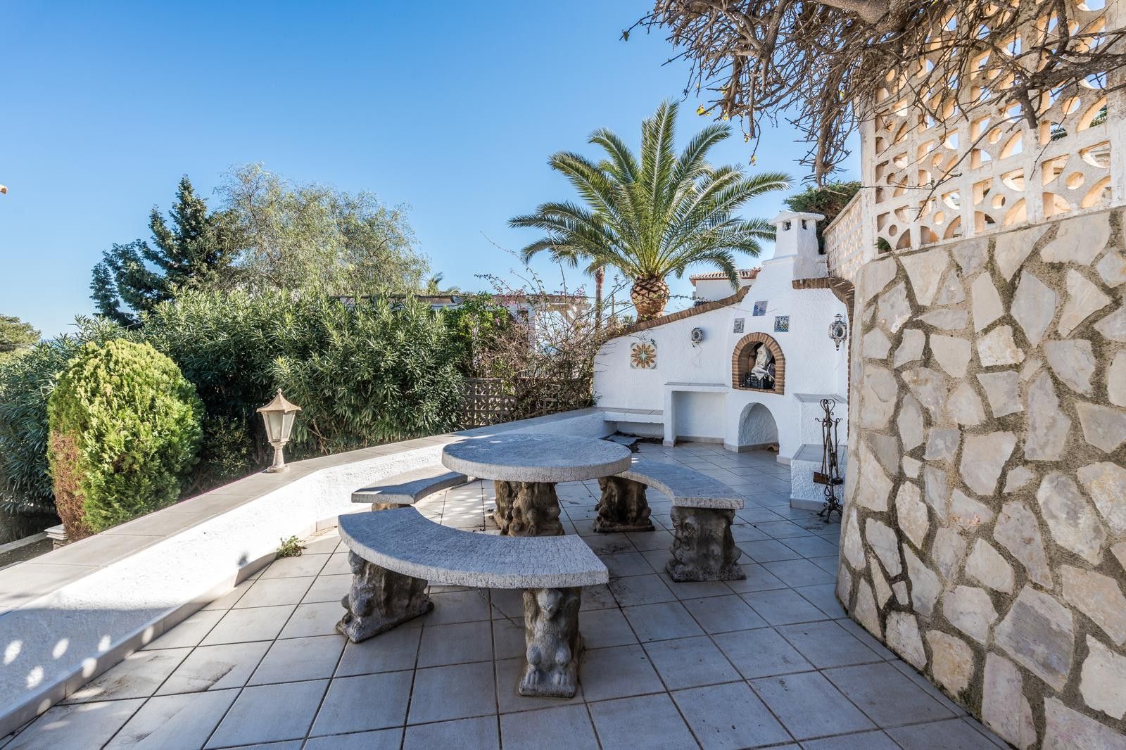 Terraza exterior con mesa y bancos de piedra tallada, zona de barbacoa de obra blanca y muros de piedra natural rodeados de vegetación mediterránea.