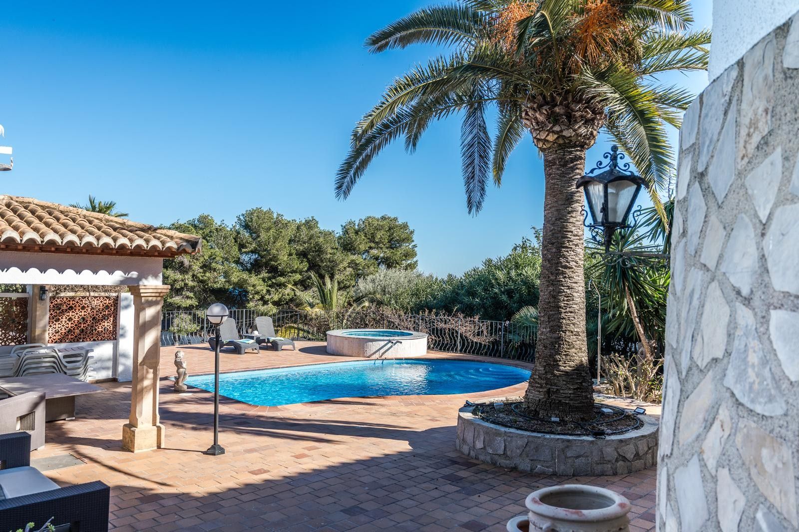Terraza exterior con piscina, jacuzzi circular y porche cubierto, rodeada de jardines mediterráneos con palmeras y suelos de terracota bajo el sol.