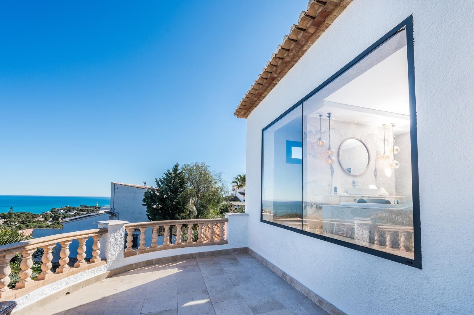 Terraza superior con balaustrada de piedra y vistas panorámicas al mar, destacando el gran ventanal que conecta el baño de diseño con el entorno mediterráneo.