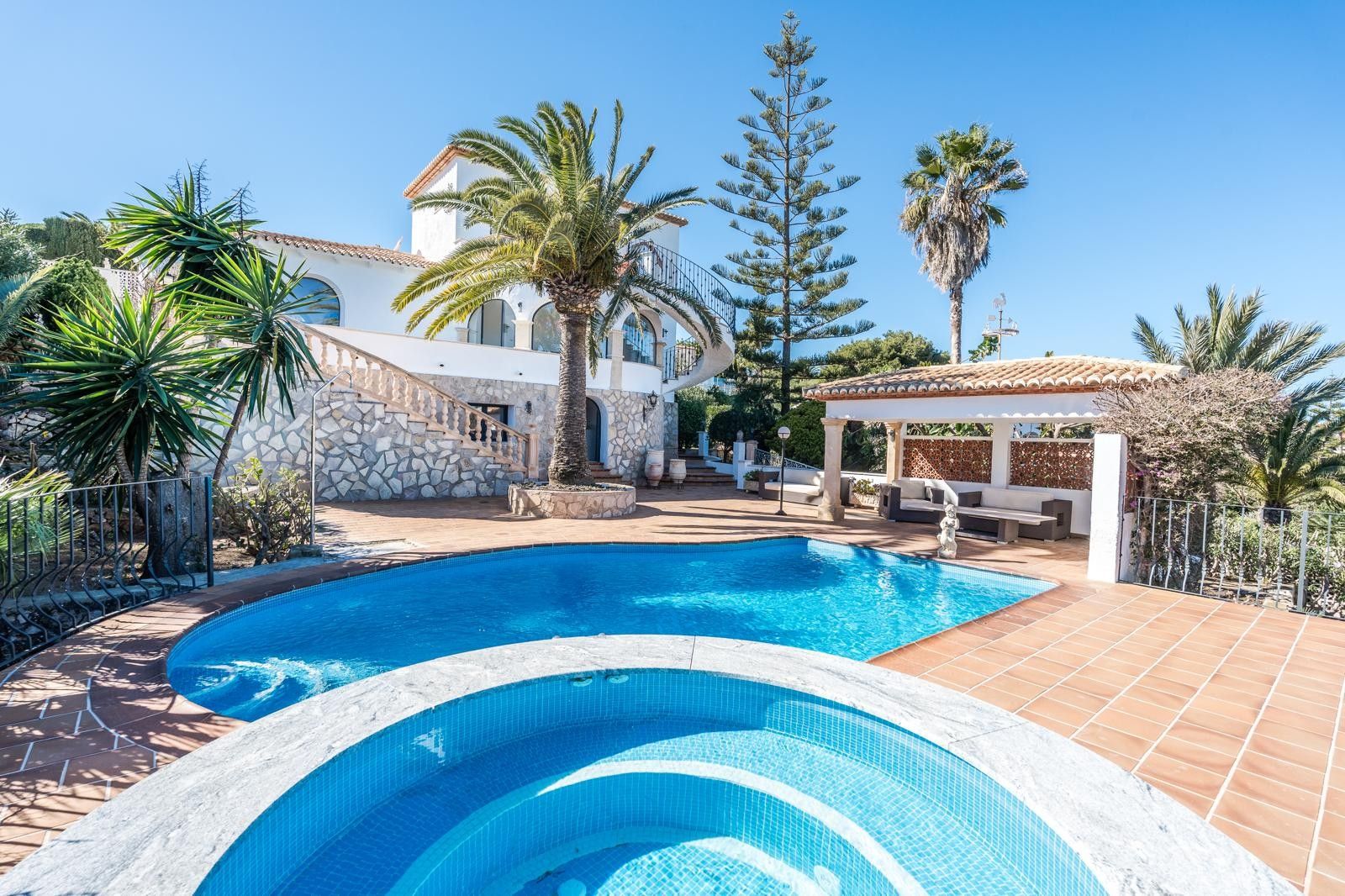 Vista exterior de la villa con piscina renovada, jacuzzi circular en primer plano y porche cubierto, rodeado de un jardín mediterráneo con palmeras y muros de piedra tallada.