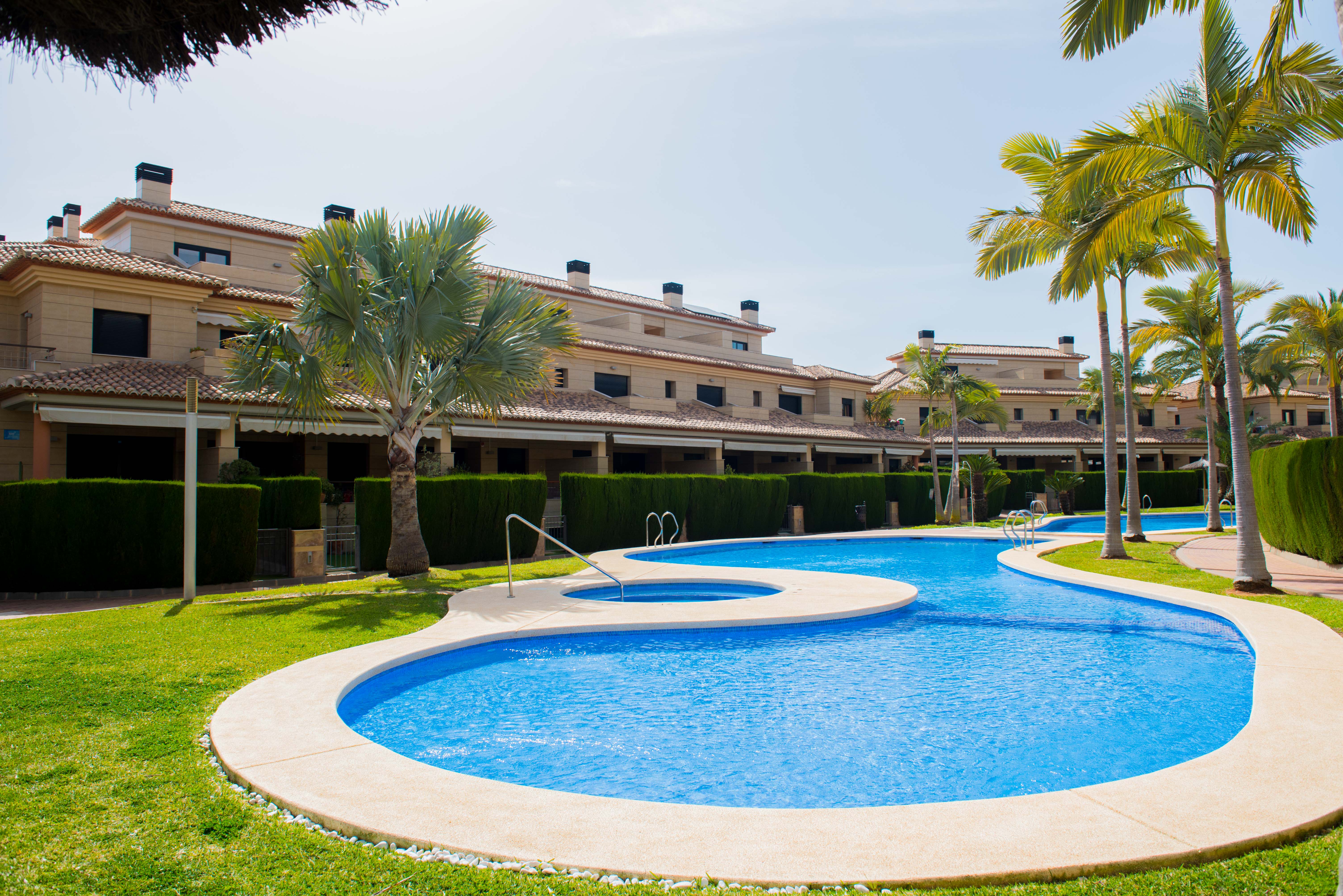 Amplia piscina comunitaria de diseño curvo con spa integrado, rodeada de césped y palmeras frente a edificios residenciales con fachadas de piedra.