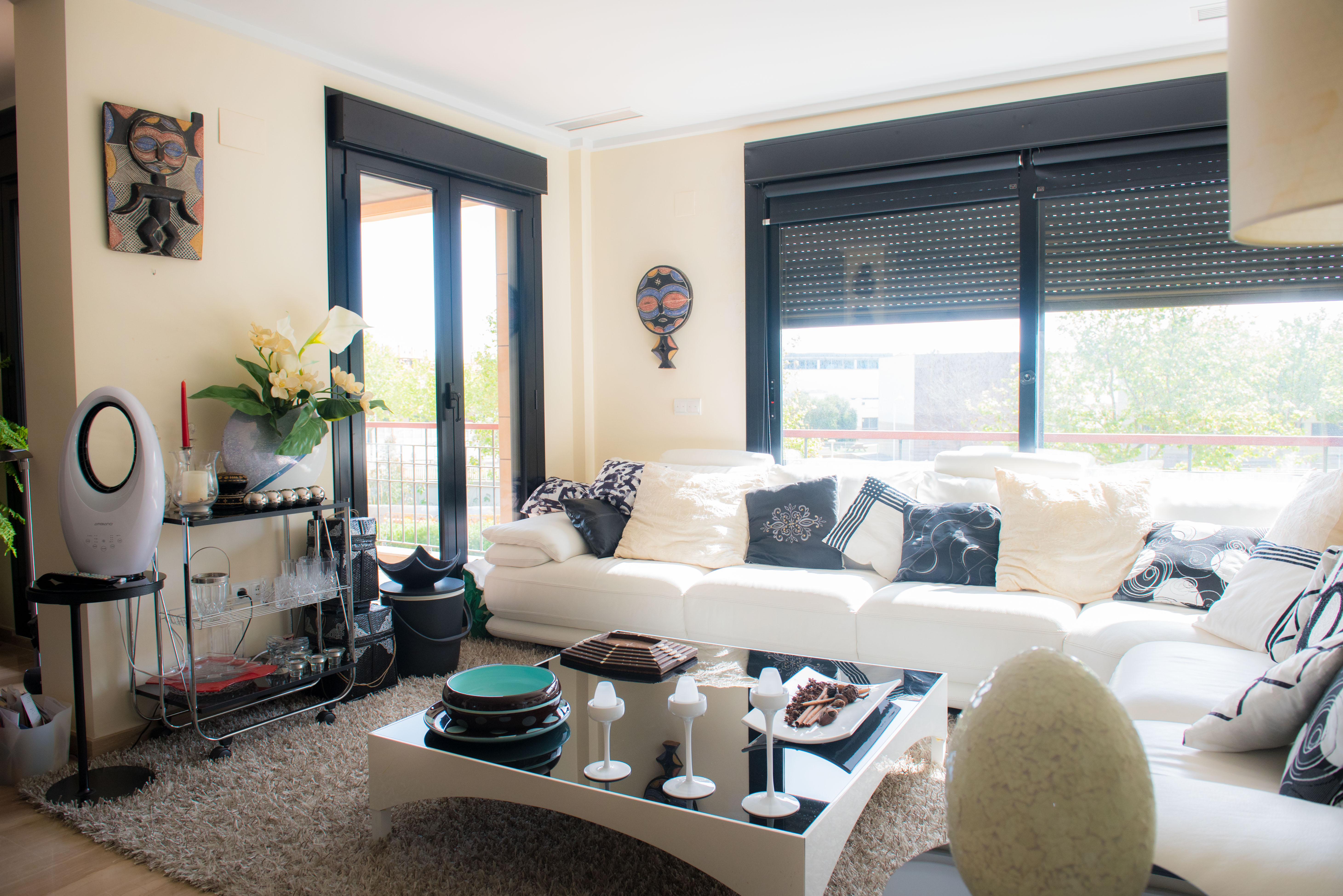 Amplio salón con sofá blanco y cojines variados, alfombra suave y mesa de centro moderna. Grandes ventanales con persianas negras y vistas a la vegetación exterior. Amplio salón con sofá blanco y cojines variados, alfombra suave y mesa de centro moderna. Grandes ventanales con persianas negras y vistas a la vegetación exterior.