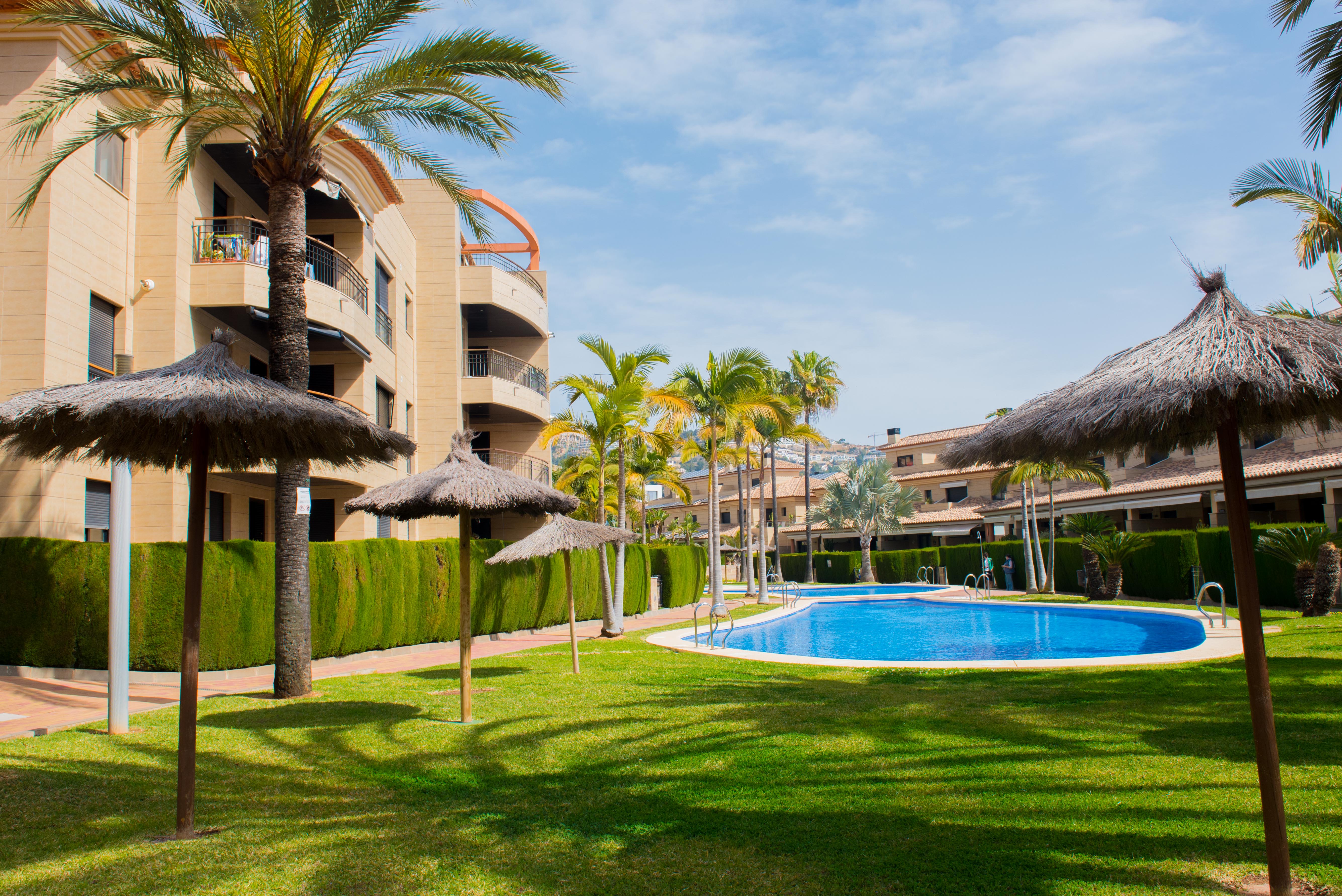 Amplios jardines comunitarios con dos piscinas, rodeadas de césped verde, palmeras y sombrillas de paja. Edificios residenciales con balcones al fondo. Amplios jardines comunitarios con dos piscinas, rodeadas de césped verde, palmeras y sombrillas de paja. Edificios residenciales con balcones al fondo.