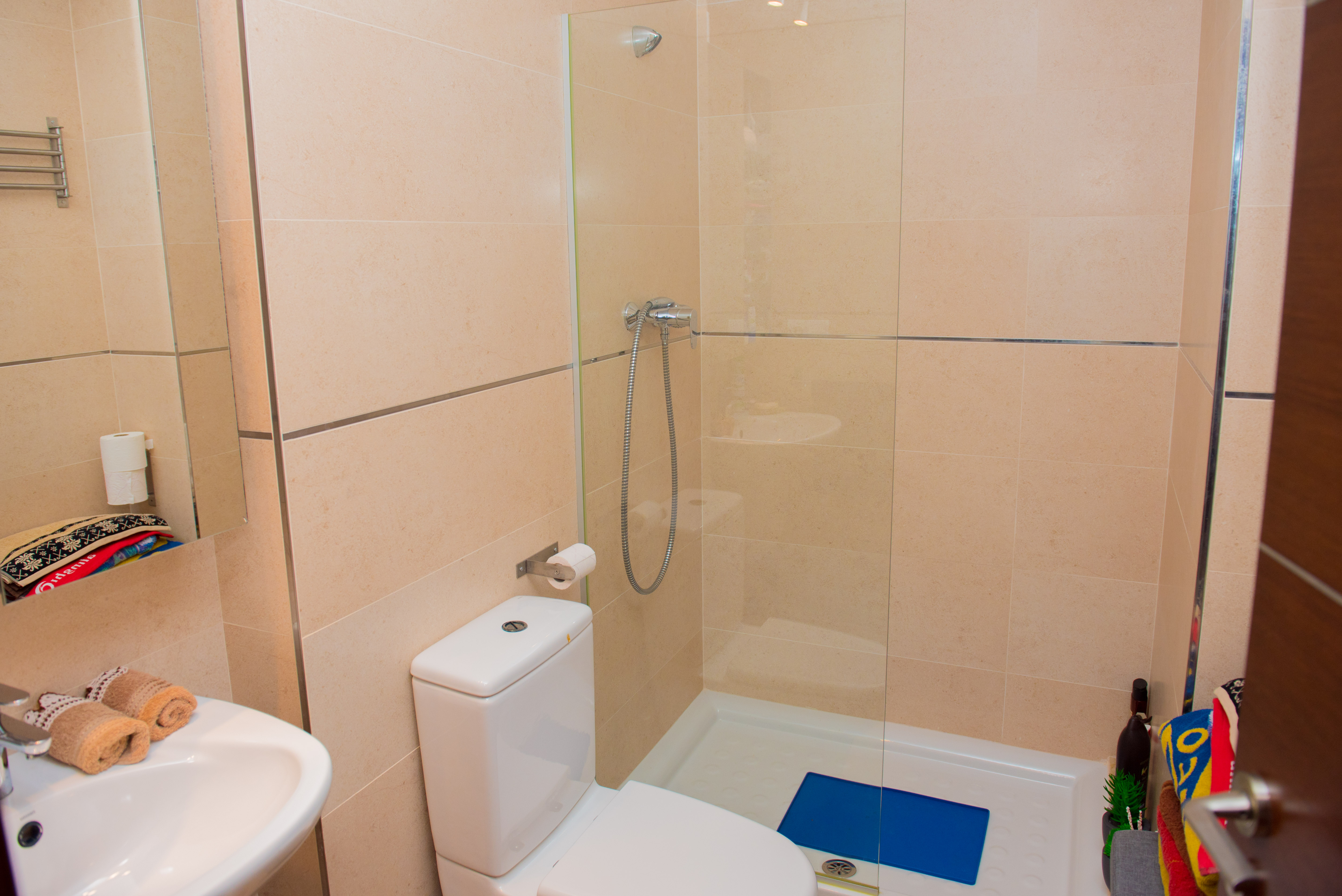 Baño moderno con ducha de obra, mampara de cristal fija, inodoro blanco y azulejos en tonos crema con perfiles metálicos decorativos.