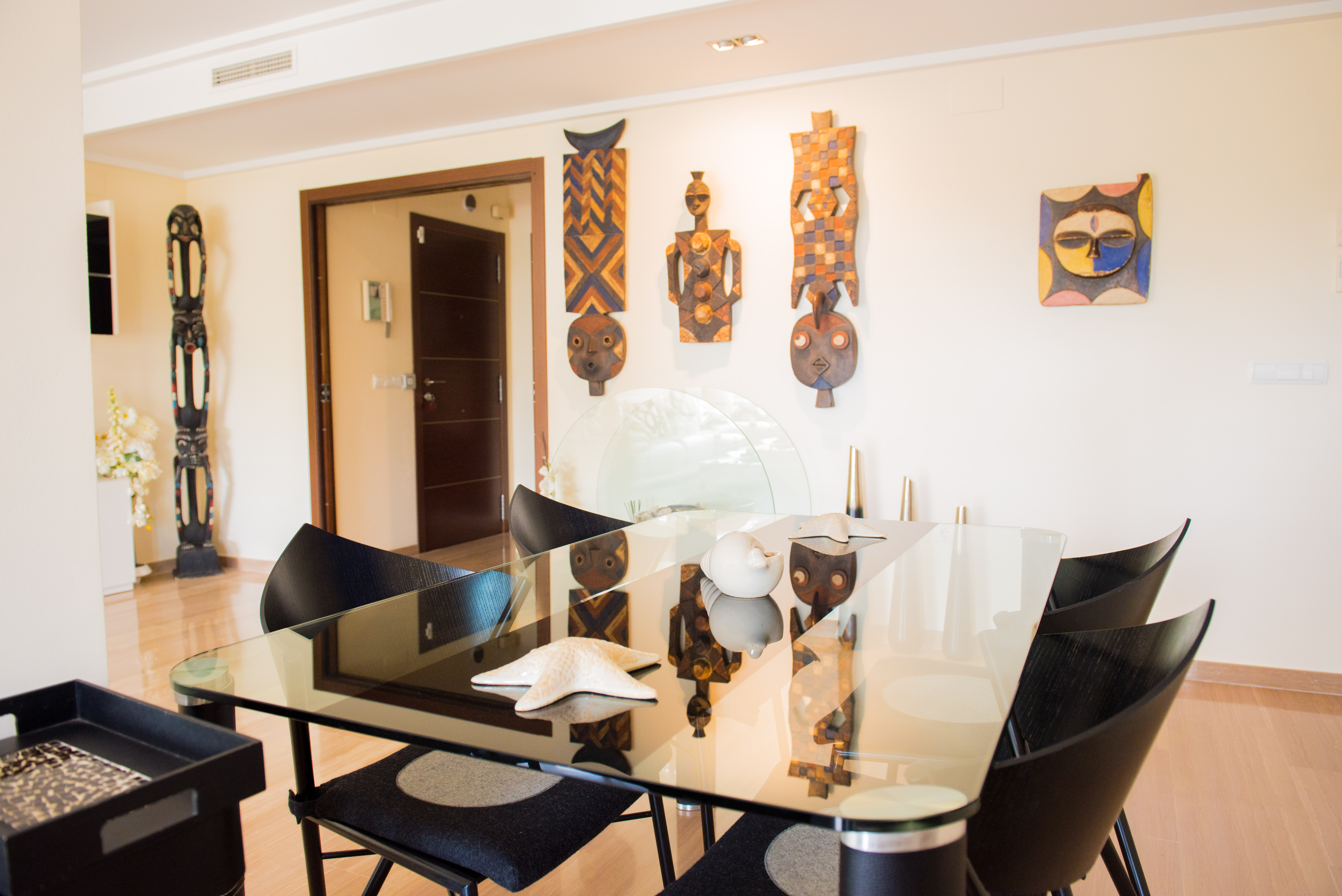Comedor contemporáneo con mesa de cristal, sillas de diseño negras y suelo de madera clara. Destacan máscaras étnicas decorativas en la pared. Comedor contemporáneo con mesa de cristal, sillas de diseño negras y suelo de madera clara. Destacan máscaras étnicas decorativas en la pared.