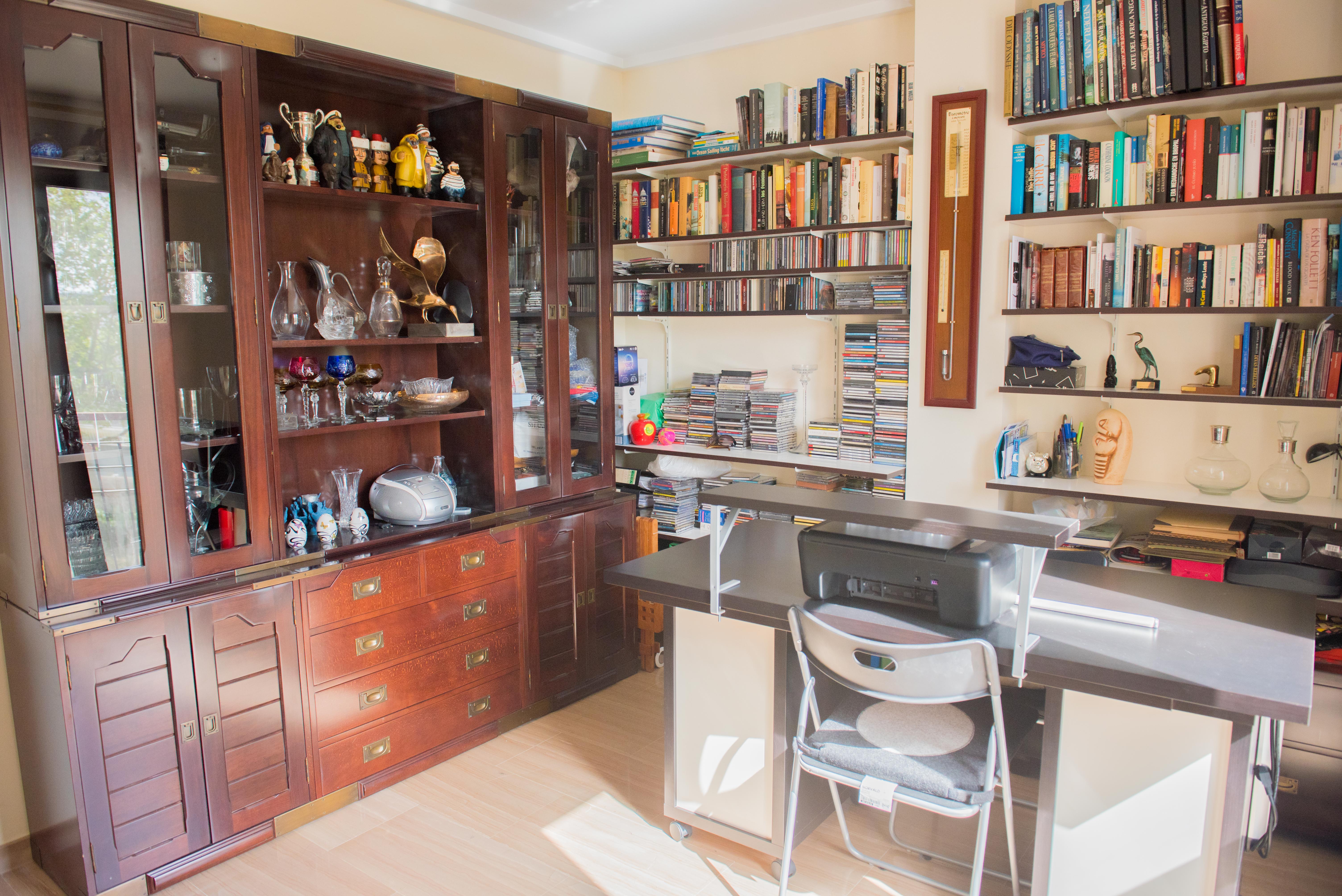Despacho con gran vitrina de madera oscura, estanterías de pared repletas de libros y zona de escritorio funcional con suelos de gres claros y luz natural.