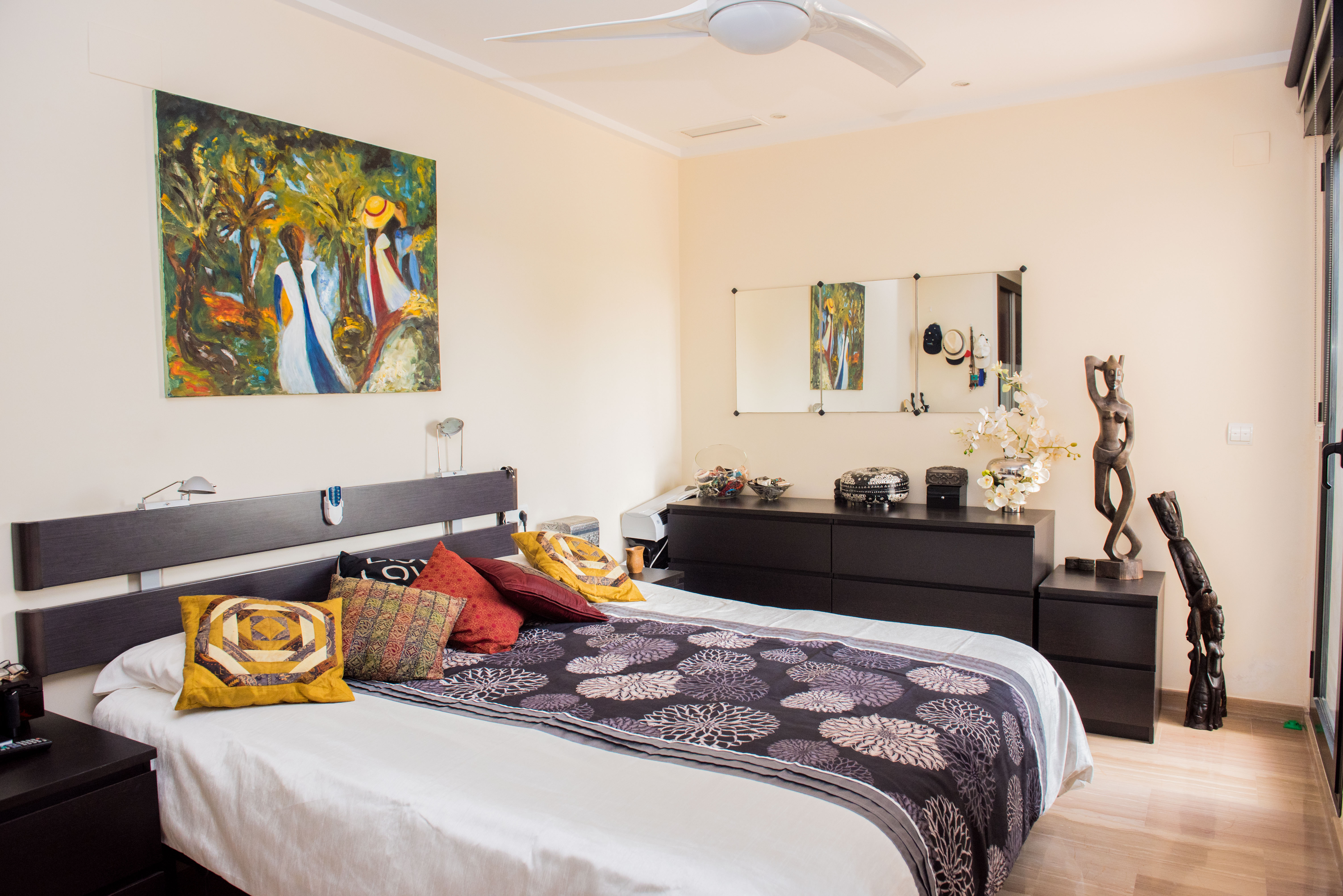 Dormitorio principal con cama doble, cabecero oscuro y mesitas a juego. Destaca una pintura abstracta sobre la cama, cómoda con espejos y ventilador de techo. Suelo de aspecto madera clara. Dormitorio principal con cama doble, cabecero oscuro y mesitas a juego. Destaca una pintura abstracta sobre la cama, cómoda con espejos y ventilador de techo. Suelo de aspecto madera clara.