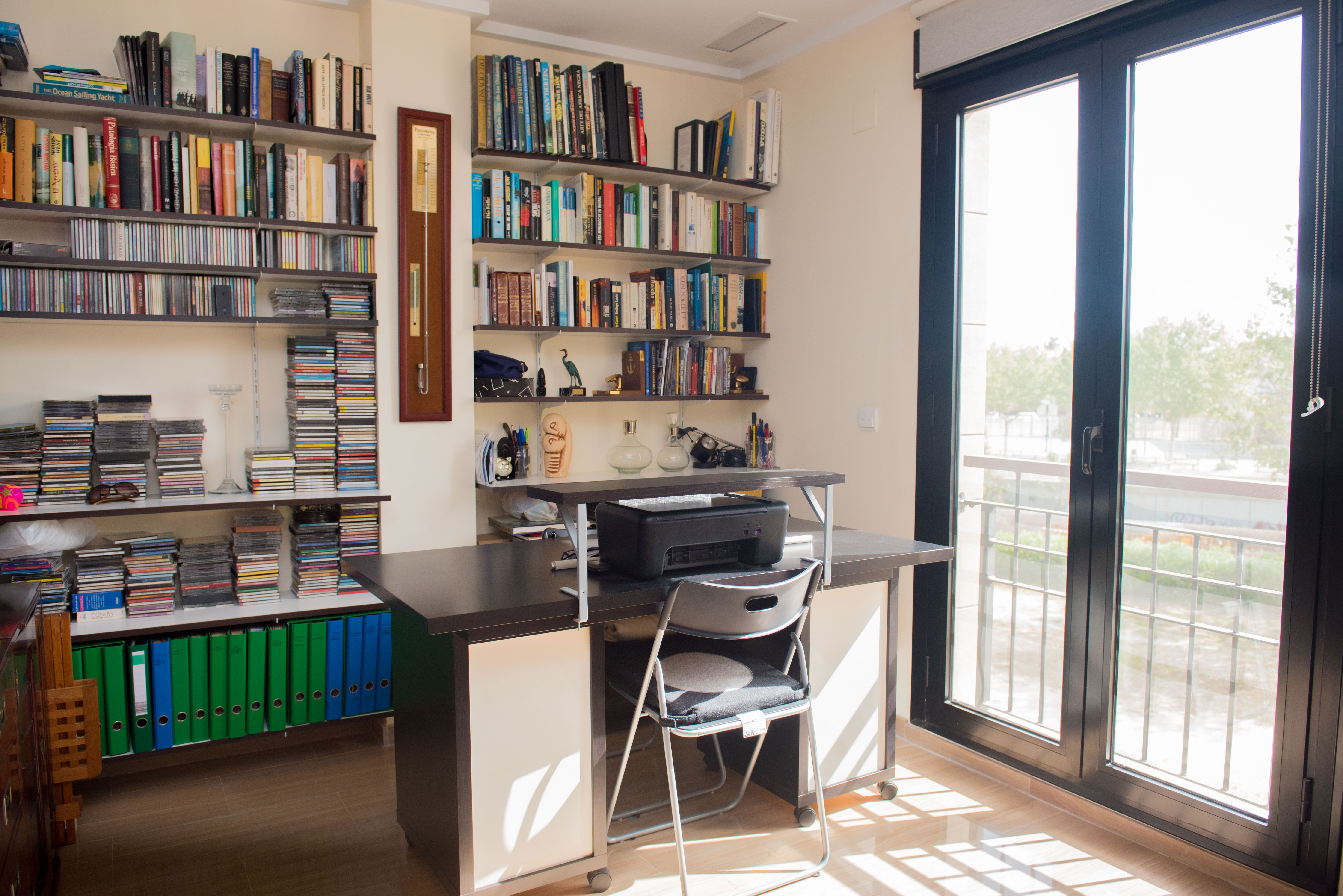 Luminoso despacho con escritorio de madera oscura, estanterías repletas de libros y CDs, y acceso directo a un balcón exterior a través de una puerta acristalada. Luminoso despacho con escritorio de madera oscura, estanterías repletas de libros y CDs, y acceso directo a un balcón exterior a través de una puerta acristalada.