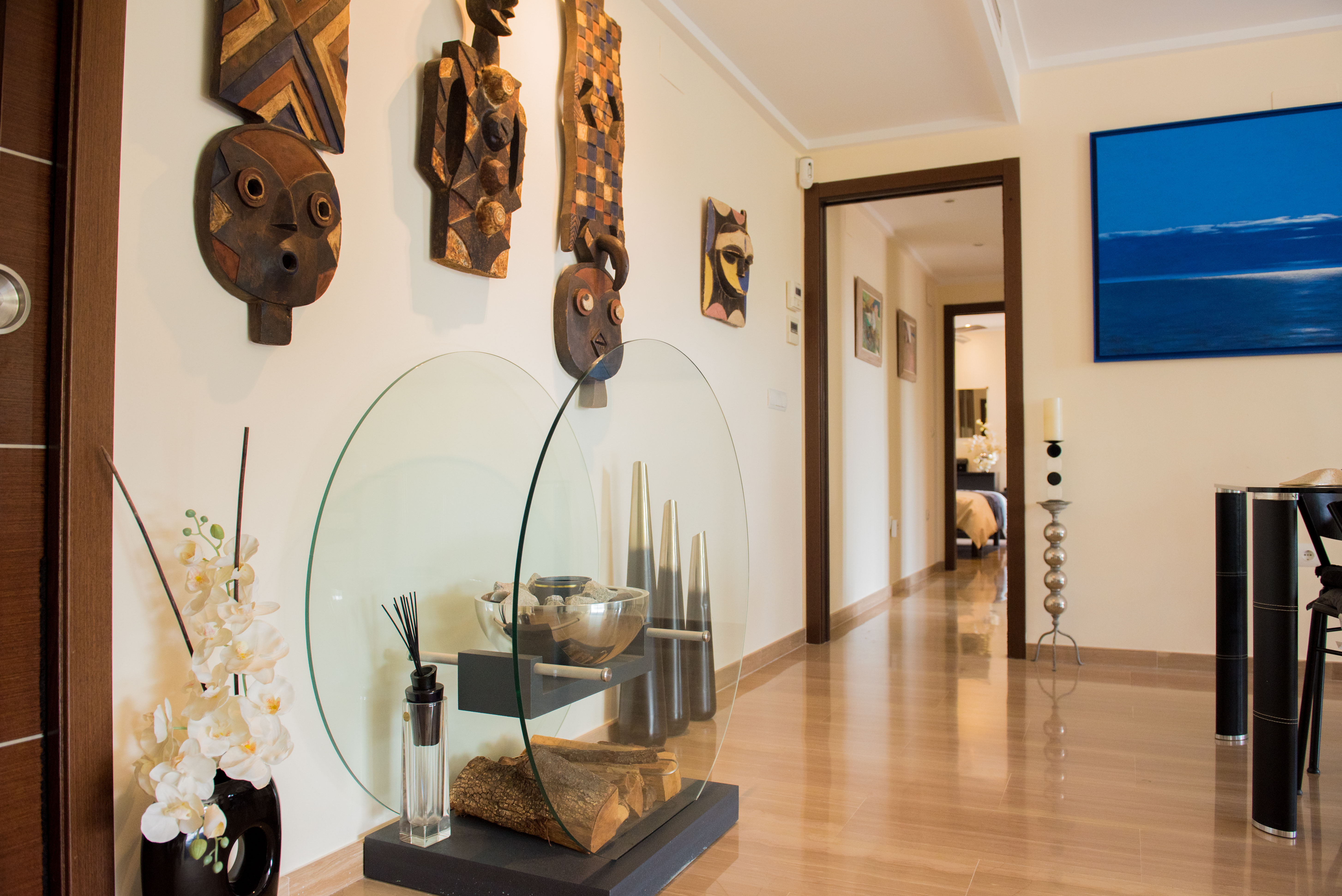 Luminoso pasillo con suelo de gres porcelánico brillante efecto madera, decorado con máscaras tribales de madera y un moderno expositor de cristal con leña y orquídeas. Un cuadro abstracto azul completa la decoración. Luminoso pasillo con suelo de gres porcelánico brillante efecto madera, decorado con máscaras tribales de madera y un moderno expositor de cristal con leña y orquídeas. Un cuadro abstracto azul completa la decoración.