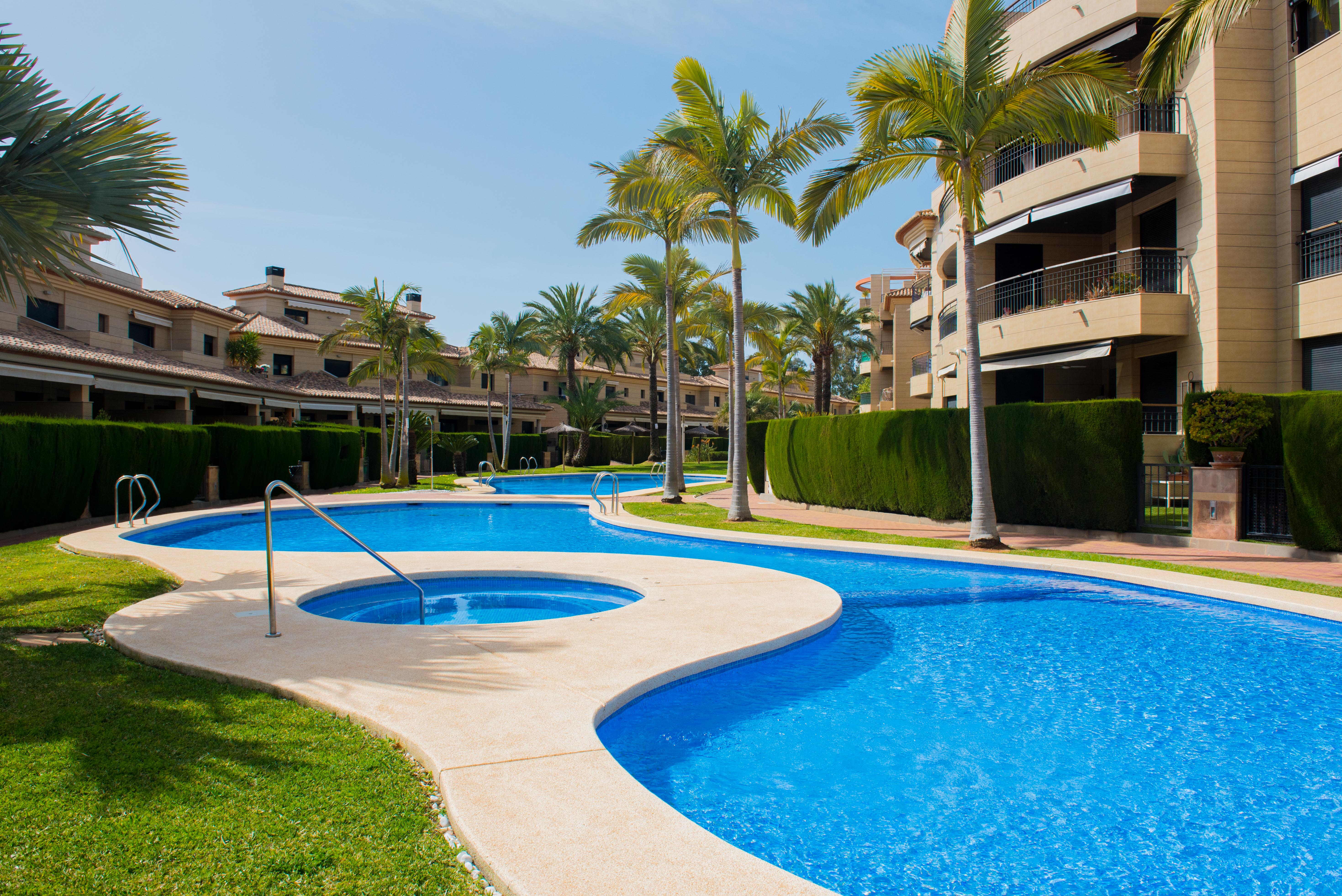 Piscina comunitaria de diseño curvo con jacuzzi integrado, rodeada de jardines mediterráneos, palmeras y edificios residenciales con fachadas de piedra clara.