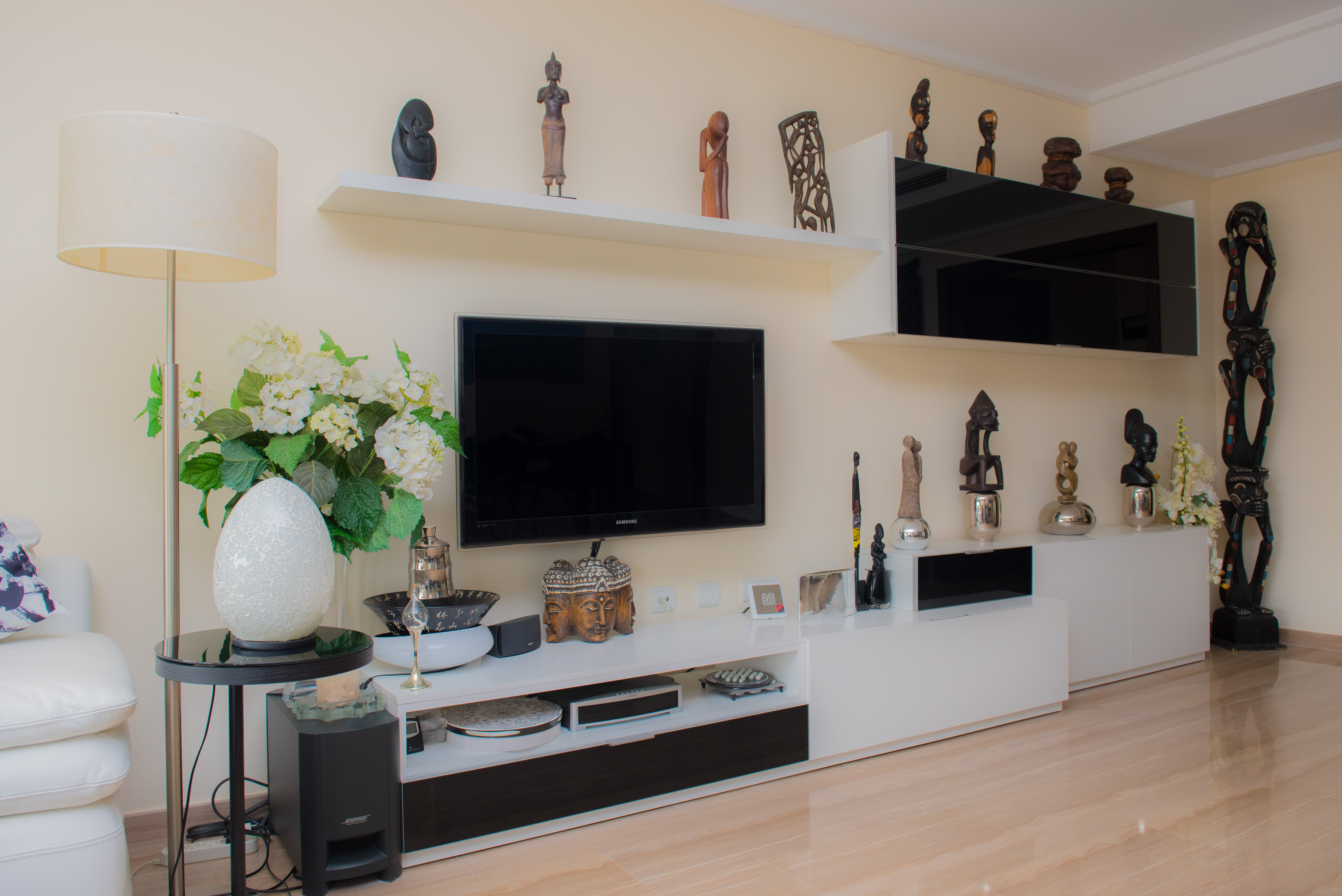 Salón con mueble de TV en blanco y negro, estanterías flotantes exhibiendo esculturas variadas y suelo de gres porcelánico claro. Incluye una lámpara de pie y detalles decorativos. Salón con mueble de TV en blanco y negro, estanterías flotantes exhibiendo esculturas variadas y suelo de gres porcelánico claro. Incluye una lámpara de pie y detalles decorativos.