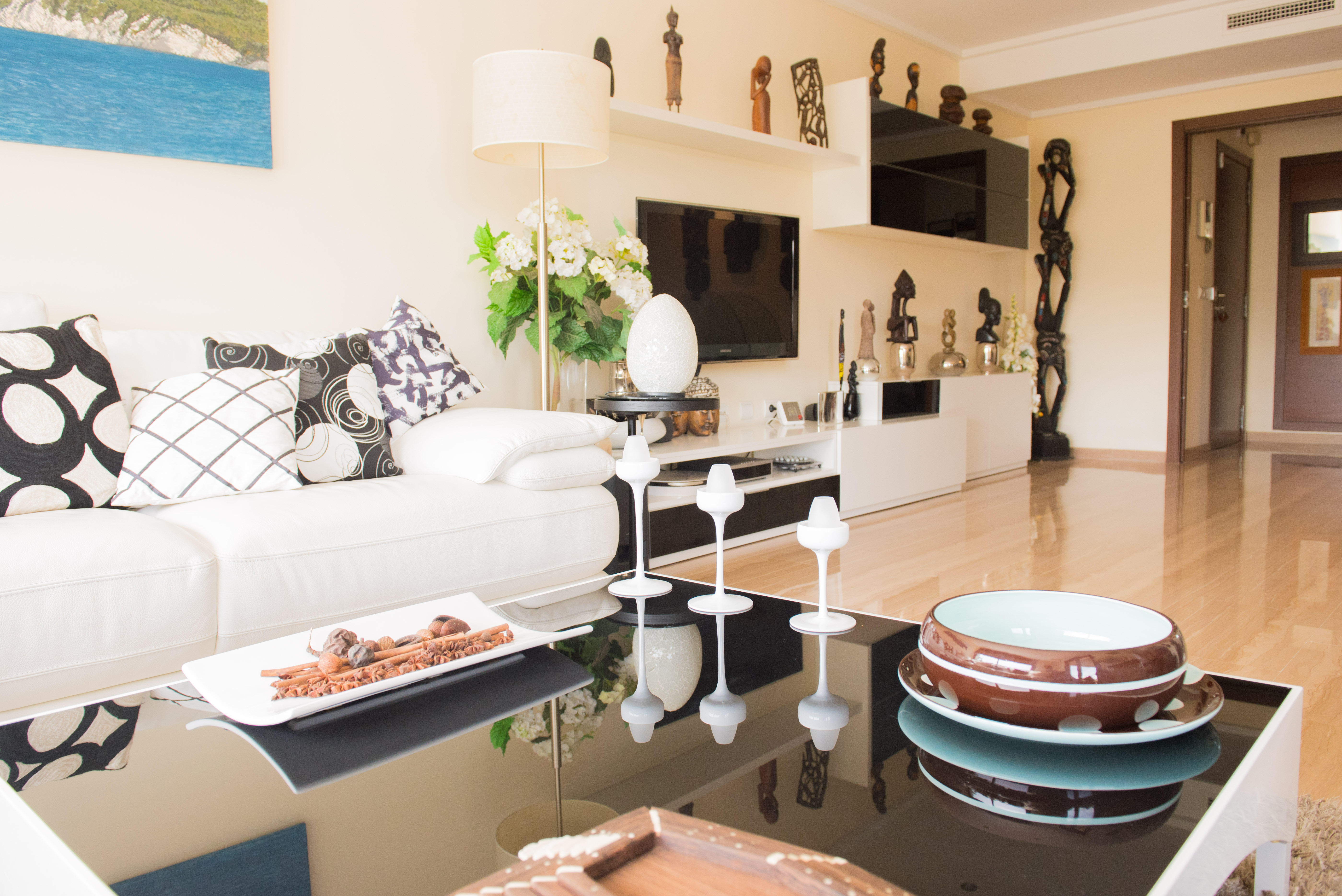 Salón con sofá blanco, cojines gráficos y mesa de centro negra brillante con detalles decorativos. Mueble de TV con estantes y esculturas africanas. Salón con sofá blanco, cojines gráficos y mesa de centro negra brillante con detalles decorativos. Mueble de TV con estantes y esculturas africanas.