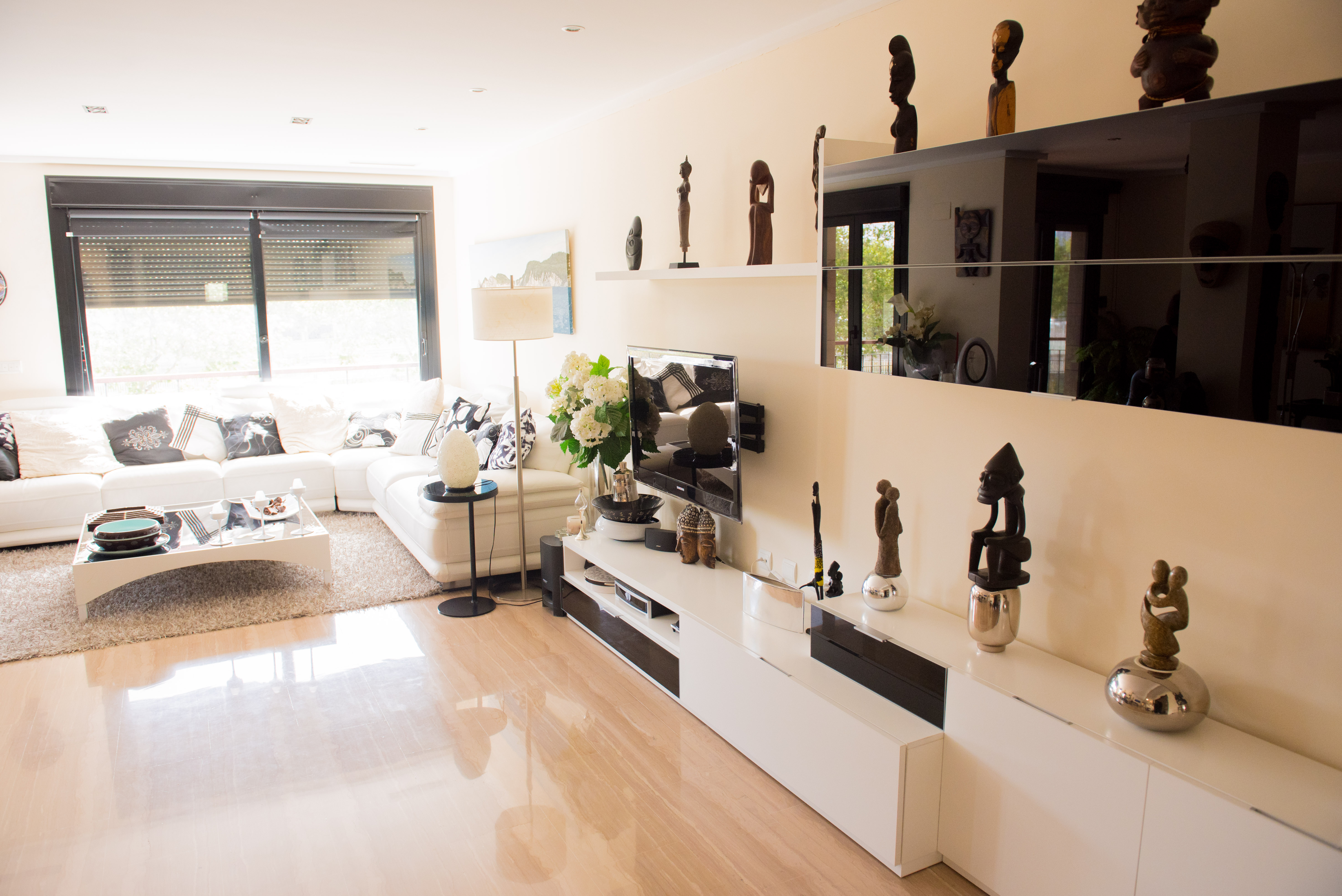 Salón luminoso con sofá seccional blanco, suelo de baldosas pulidas y mueble de TV moderno. La luz natural entra por la ventana con persianas enrollables. Salón luminoso con sofá seccional blanco, suelo de baldosas pulidas y mueble de TV moderno. La luz natural entra por la ventana con persianas enrollables.