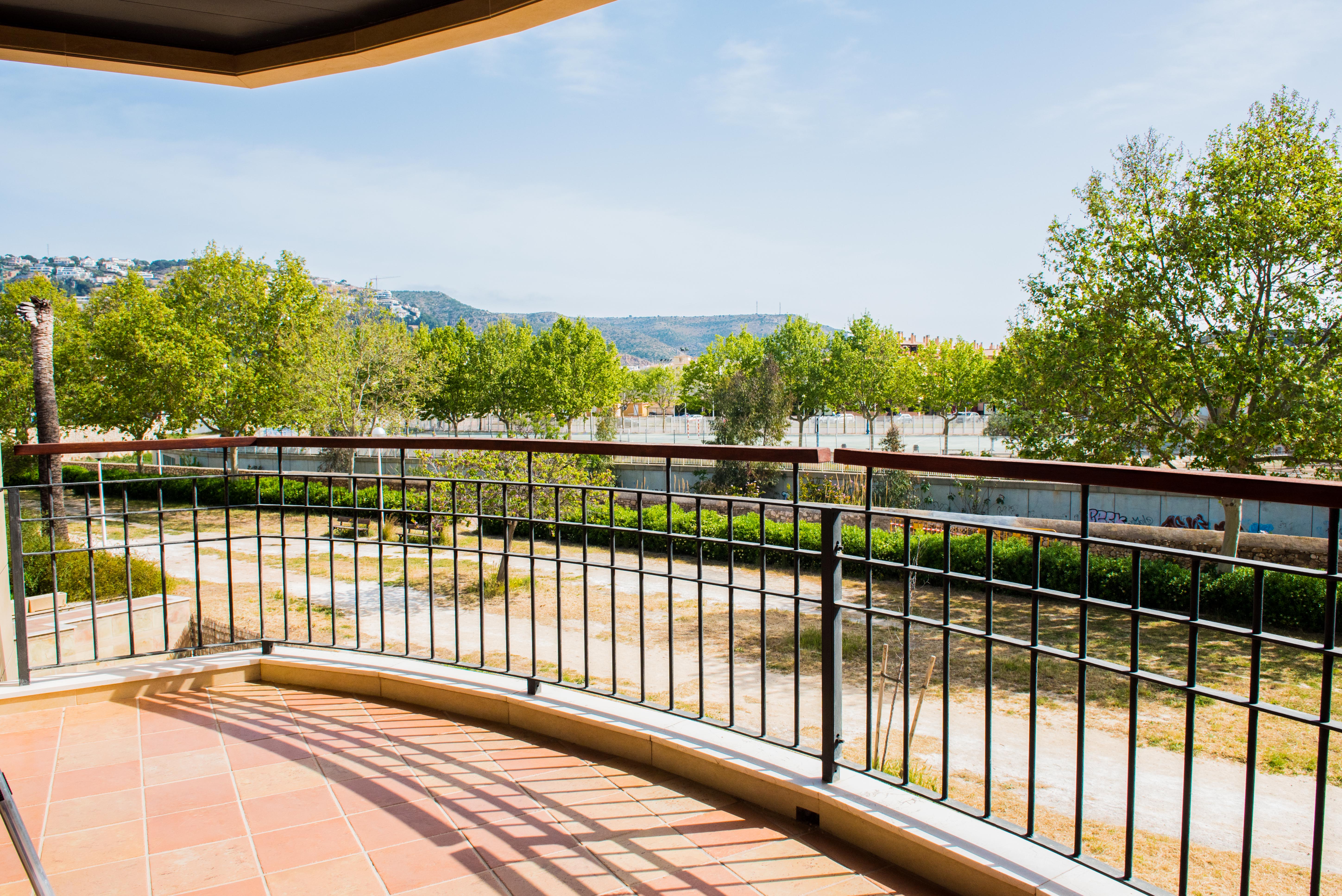 Terraza cubierta con suelo de terracota y barandilla de hierro con pasamanos de madera, con vistas despejadas a zonas verdes y montañas de Jávea.