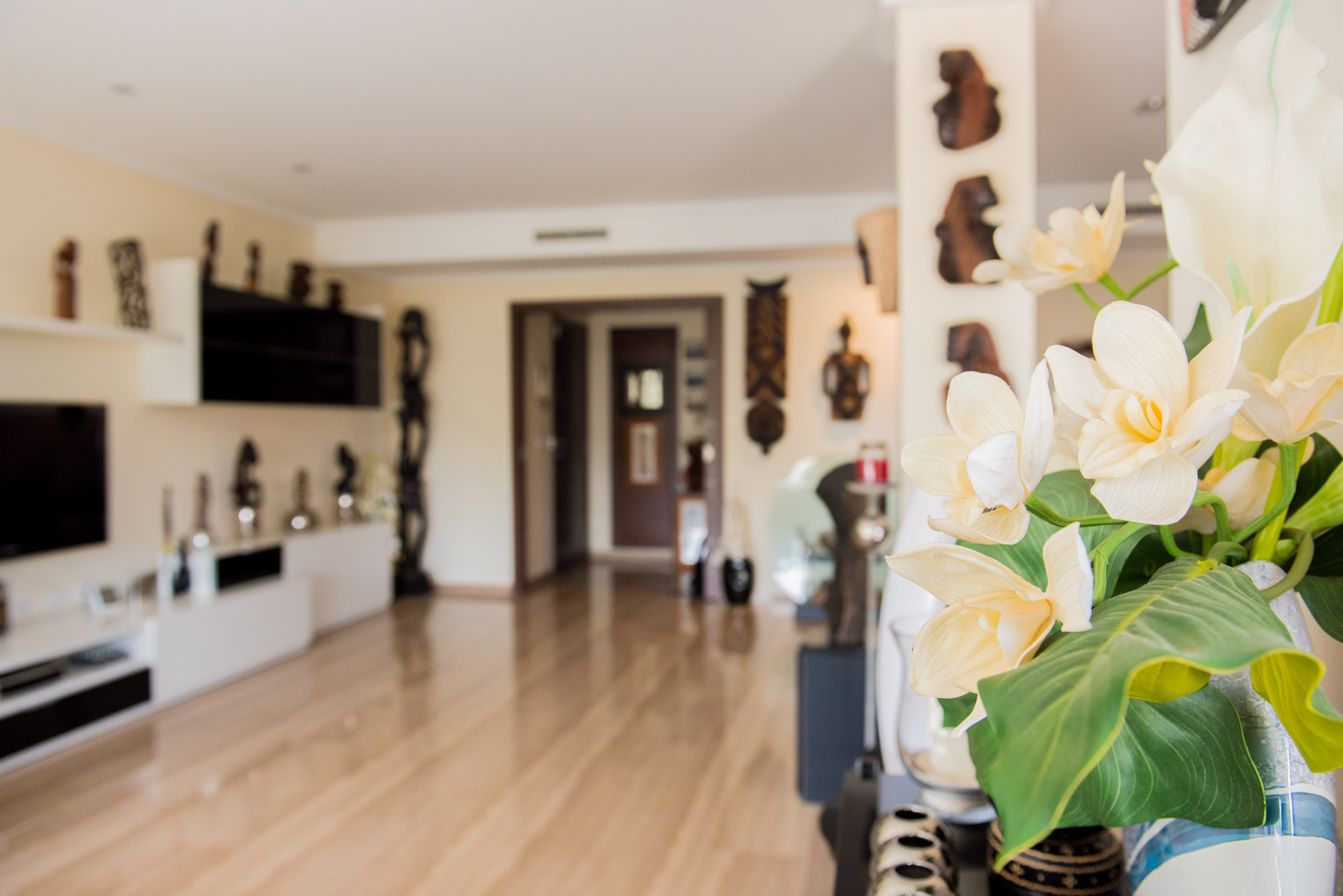 Vaso en primer plano con elegantes flores artificiales blancas y amarillas. Interior luminoso con suelo de madera clara, que conduce a una moderna sala de estar con arte mural decorativo. Vaso en primer plano con elegantes flores artificiales blancas y amarillas. Interior luminoso con suelo de madera clara, que conduce a una moderna sala de estar con arte mural decorativo.