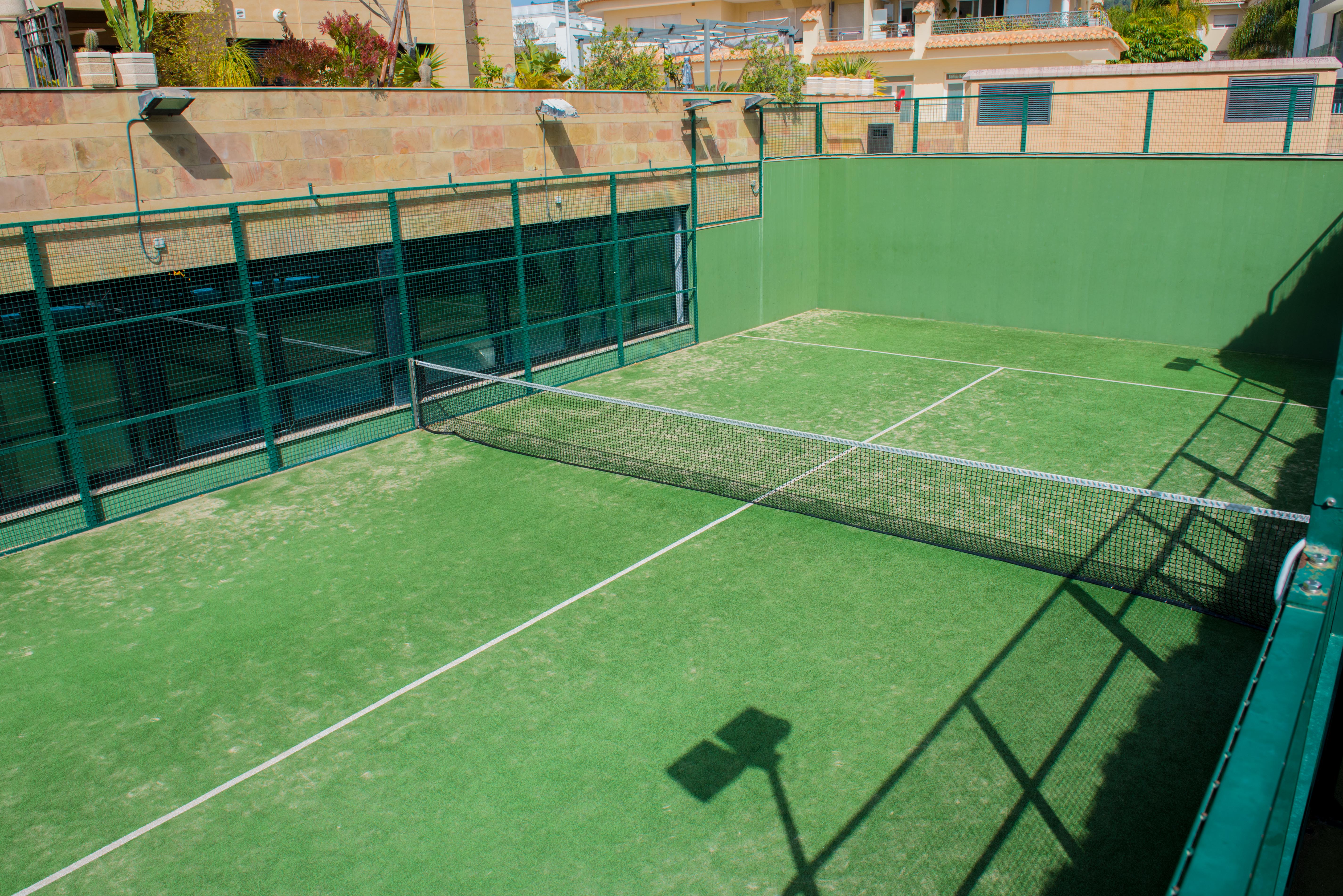 Vista de la pista de pádel exterior de la comunidad, con césped artificial verde vibrante, red y robustas paredes de malla verde, y edificios residenciales al fondo soleado. Vista de la pista de pádel exterior de la comunidad, con césped artificial verde vibrante, red y robustas paredes de malla verde, y edificios residenciales al fondo soleado.