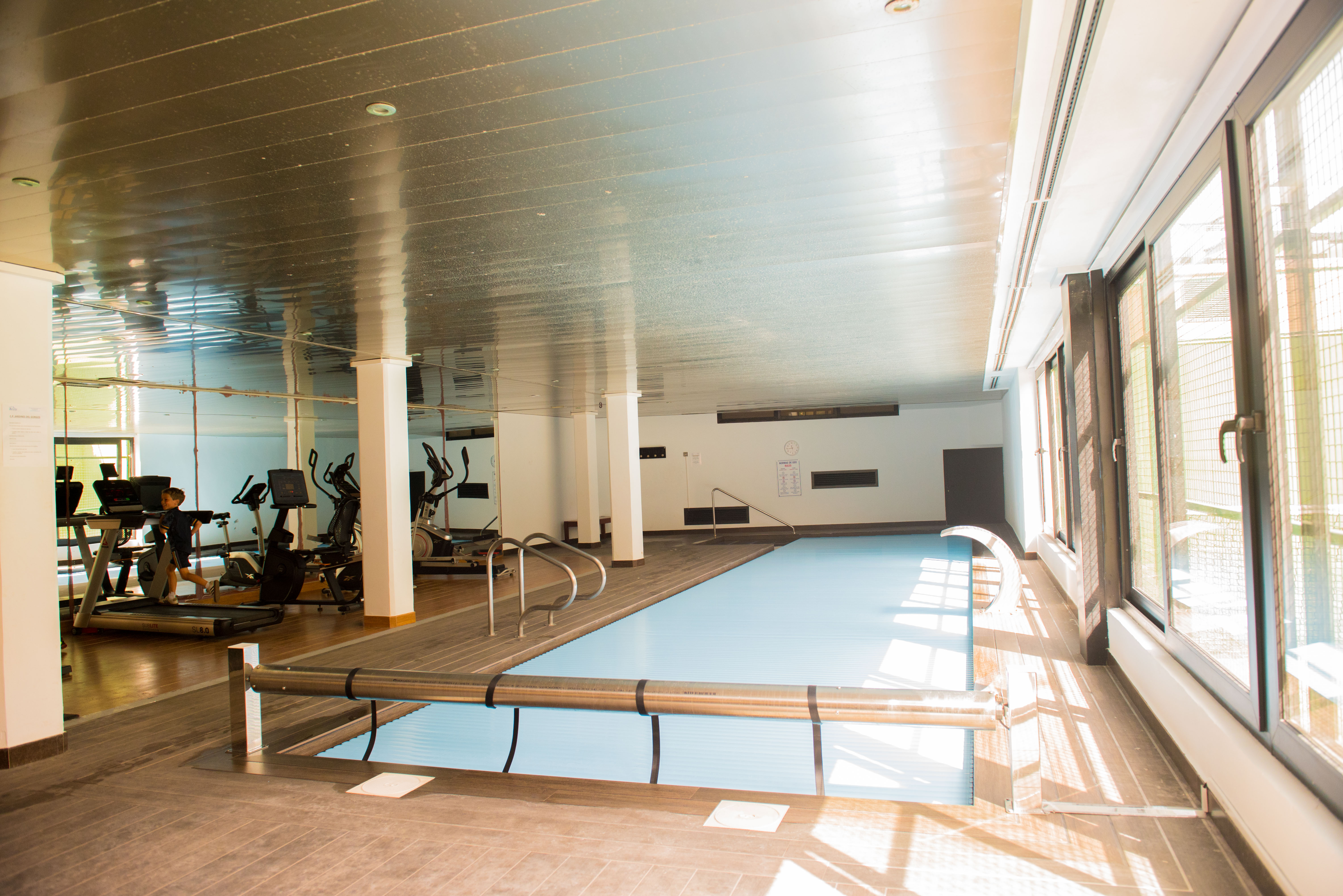 Zona de bienestar interior con piscina climatizada cubierta y gimnasio equipado con maquinaria de cardio, iluminada por amplios ventanales laterales.