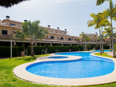 Amplia piscina comunitaria de forma orgánica con jacuzzi integrado, rodeada de césped verde y palmeras, frente a edificios residenciales con tejados de teja.
