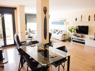 Amplio salón-comedor de diseño contemporáneo con suelo de madera clara. Destaca la mesa de cristal con sillas negras, el sofá blanco y el acceso a una terraza a través de puertas acristaladas oscuras.