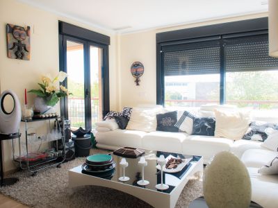 Amplio salón con sofá blanco y cojines variados, alfombra suave y mesa de centro moderna. Grandes ventanales con persianas negras y vistas a la vegetación exterior.