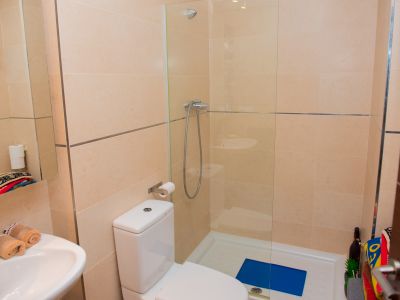Baño moderno con paredes de azulejos beige claro y detalles metálicos. Incluye inodoro blanco, lavabo con grifo cromado y ducha con mampara de cristal y plato blanco.
