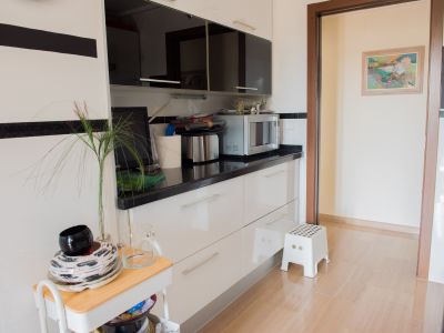 Cocina moderna con mobiliario blanco y encimera negra, equipada con microondas. El suelo de baldosas claras y el acceso a otra estancia con un cuadro complementan el diseño funcional.