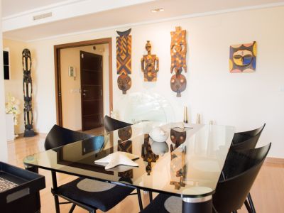 Comedor contemporáneo con mesa de cristal, sillas de diseño negras y suelo de madera clara. Destacan máscaras étnicas decorativas en la pared.