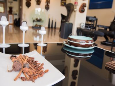 Detalle de una mesa reflectante con una bandeja de especias (canela, anís estrellado), modernos candelabros blancos y cuencos cerámicos. El fondo muestra un interior decorado.