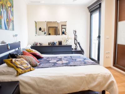 Dormitorio principal con cama doble, cabecero oscuro con lámparas de lectura y acceso a terraza. Incluye un armario empotrado de madera y cristal, suelo laminado claro y cómoda con espejos.