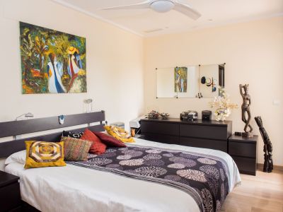 Dormitorio principal con cama doble, cabecero oscuro y mesitas a juego. Destaca una pintura abstracta sobre la cama, cómoda con espejos y ventilador de techo. Suelo de aspecto madera clara.