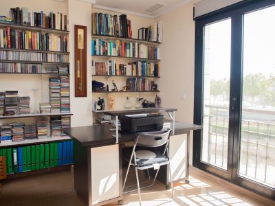 Luminoso despacho con escritorio de madera oscura, estanterías repletas de libros y CDs, y acceso directo a un balcón exterior a través de una puerta acristalada.